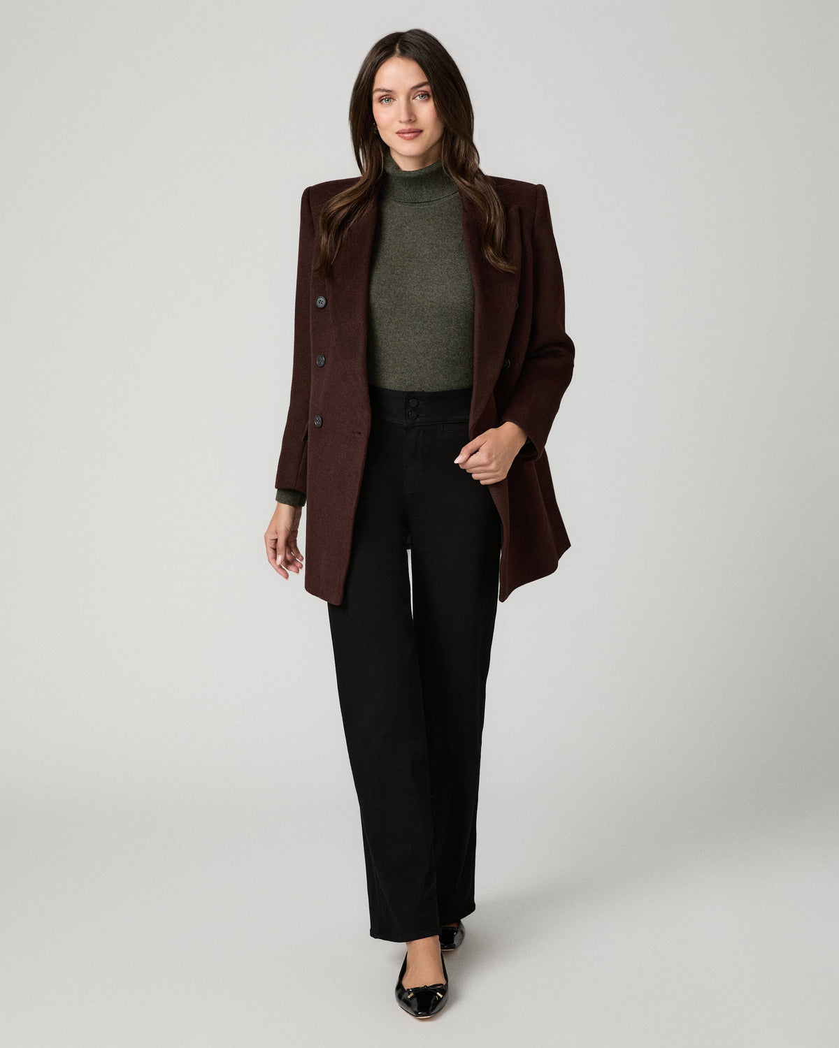 A woman modeling a dark brown structured blazer over a dark green turtleneck sweater and black wide-leg trousers, standing against a light gray studio background.