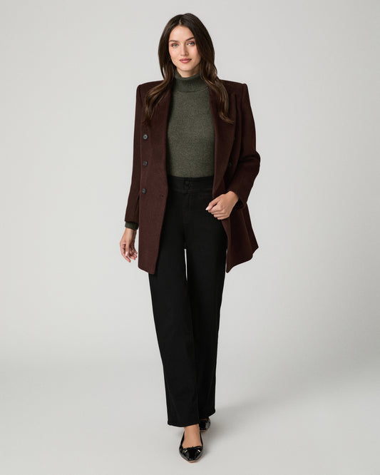 A woman modeling a dark brown structured blazer over a dark green turtleneck sweater and black wide-leg trousers, standing against a light gray studio background.