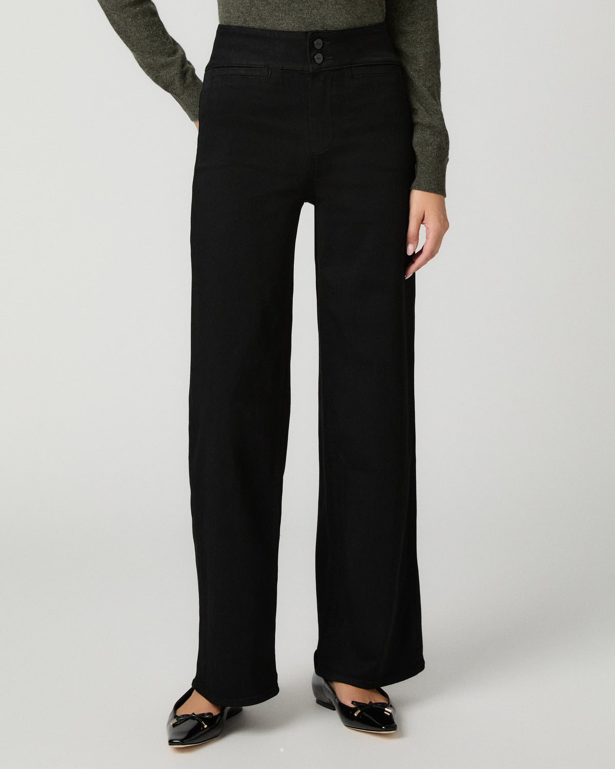 A model is wearing a pair of black wide-leg trousers with a button waistband, styled with a dark olive green long-sleeve top and black patent leather flats, shot against a light gray studio background.