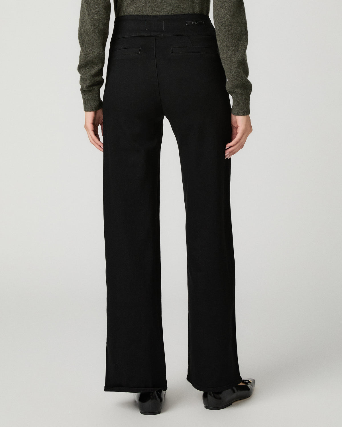 Rear view of a model wearing black wide-leg trousers with a ribbed waistband, paired with a dark green long-sleeve top and black shoes, against a light gray studio background.
