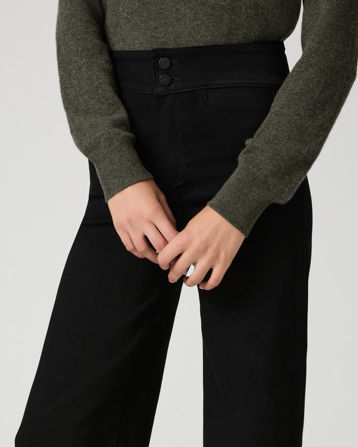 A close-up of a person wearing black wide-leg trousers with a double-button waistband and an olive green sweater, shown against a light gray studio background.