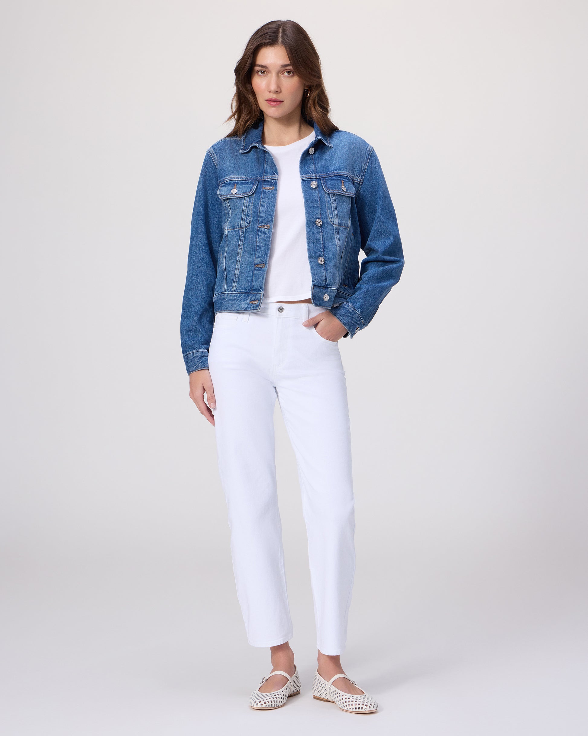 Woman wearing a blue denim jacket over a white shirt paired with white denim and white flats on a light gray background