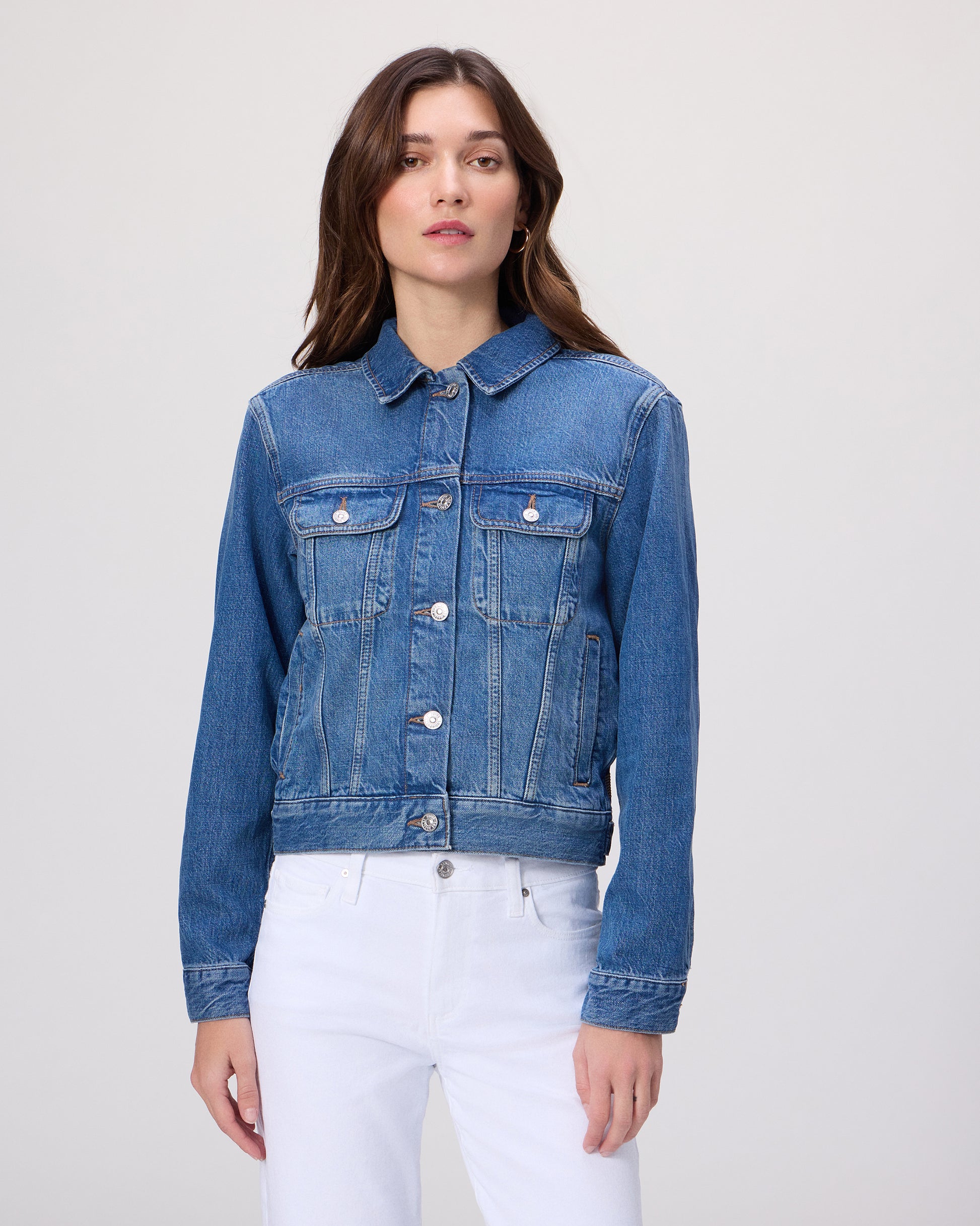 Woman wearing a blue denim jacket over a white shirt paired with white denim on a light gray background