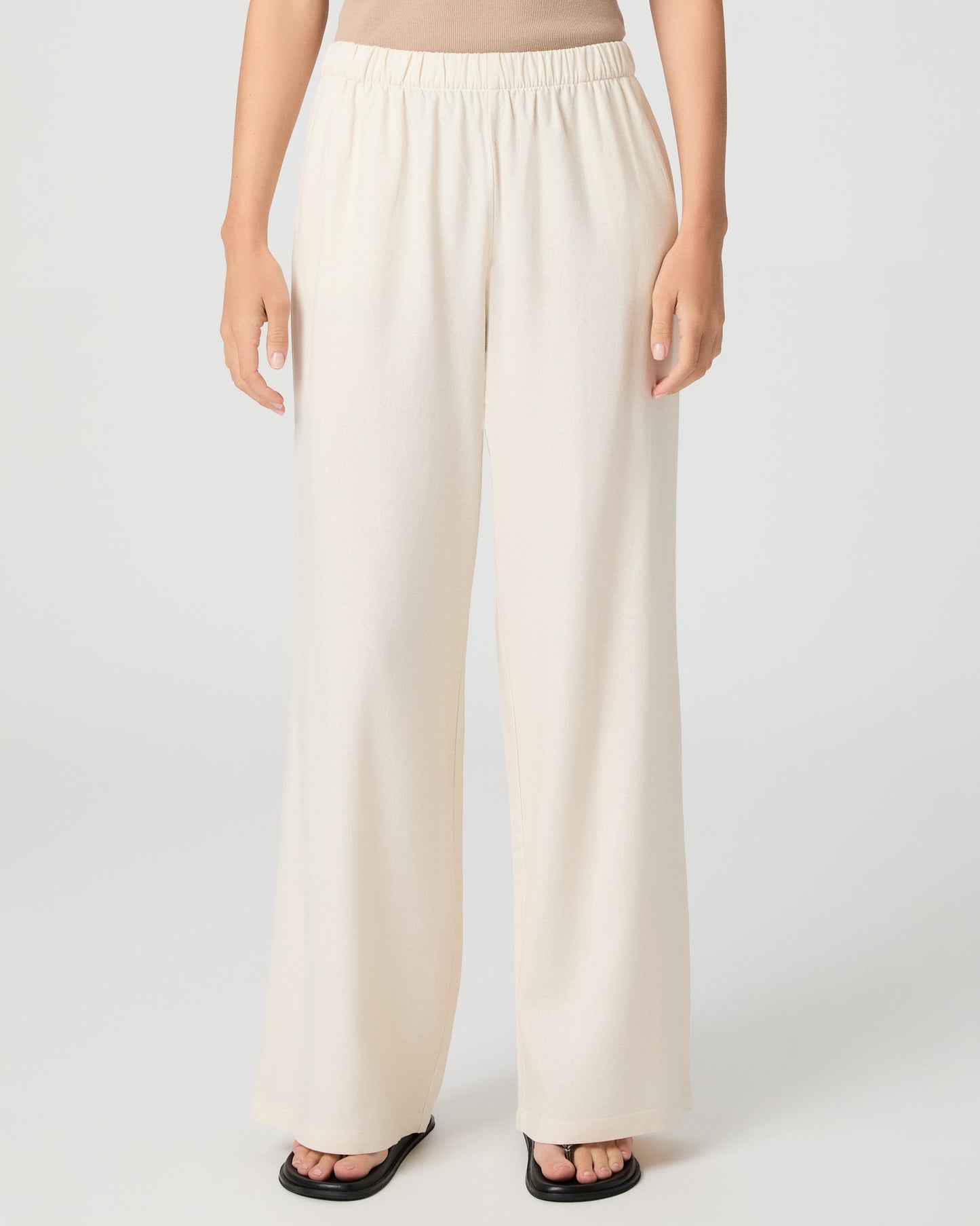 Woman wearing a beige tank top and white pants on a plain background