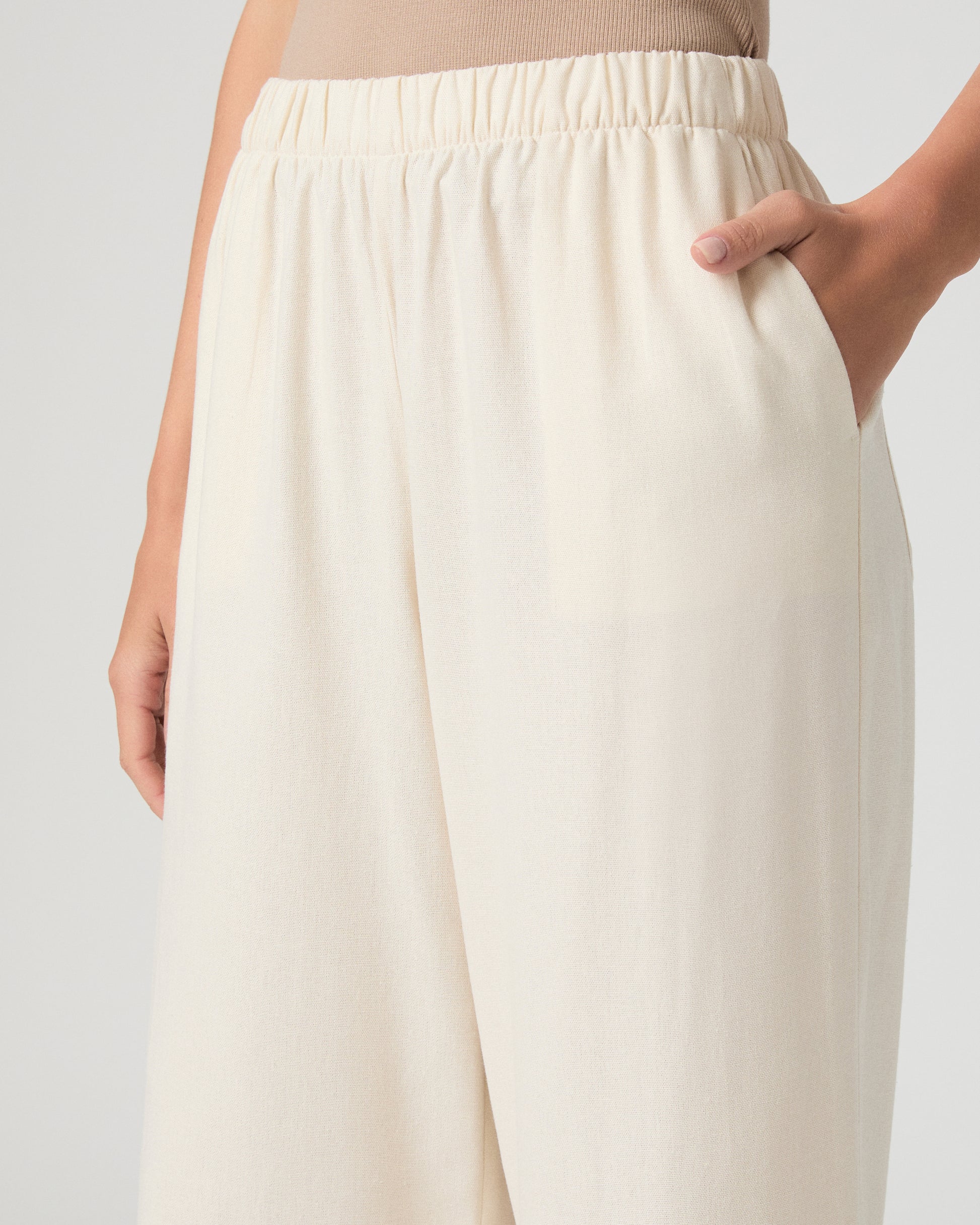 Woman wearing a beige tank top and white pants on a plain background