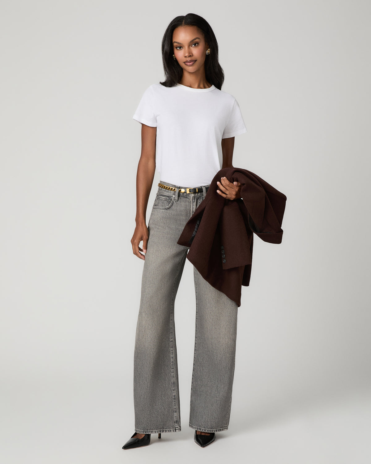 Full-length view of a woman in a white tee tucked into gray jeans with a brown belt, draping a dark brown blazer over her arm.