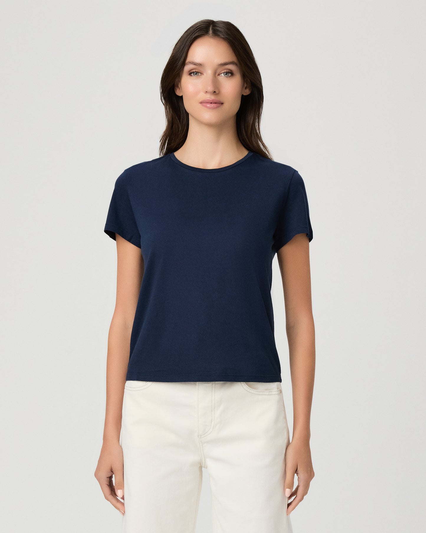 A woman modeling a navy blue short-sleeve crewneck t-shirt paired with off-white pants, shot against a solid light gray background.