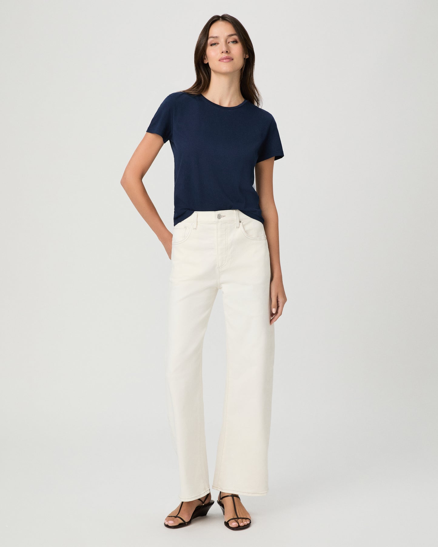A woman modeling a navy blue short-sleeve crewneck t-shirt paired with off-white pants, shot against a solid light gray background.