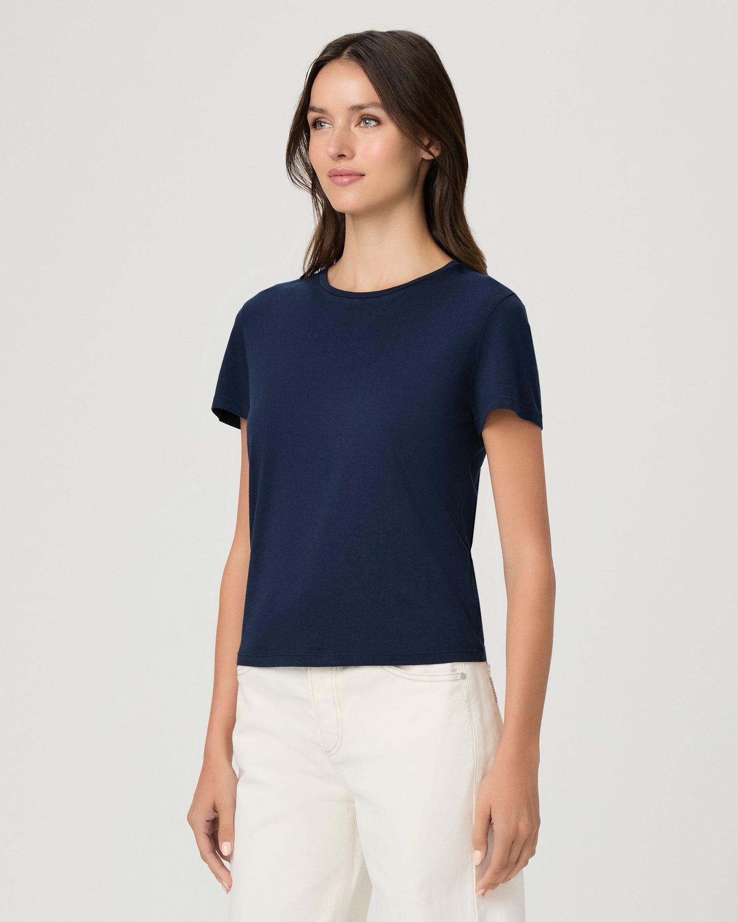 A woman modeling a navy blue short-sleeve crewneck t-shirt paired with off-white pants, shot against a solid light gray background.