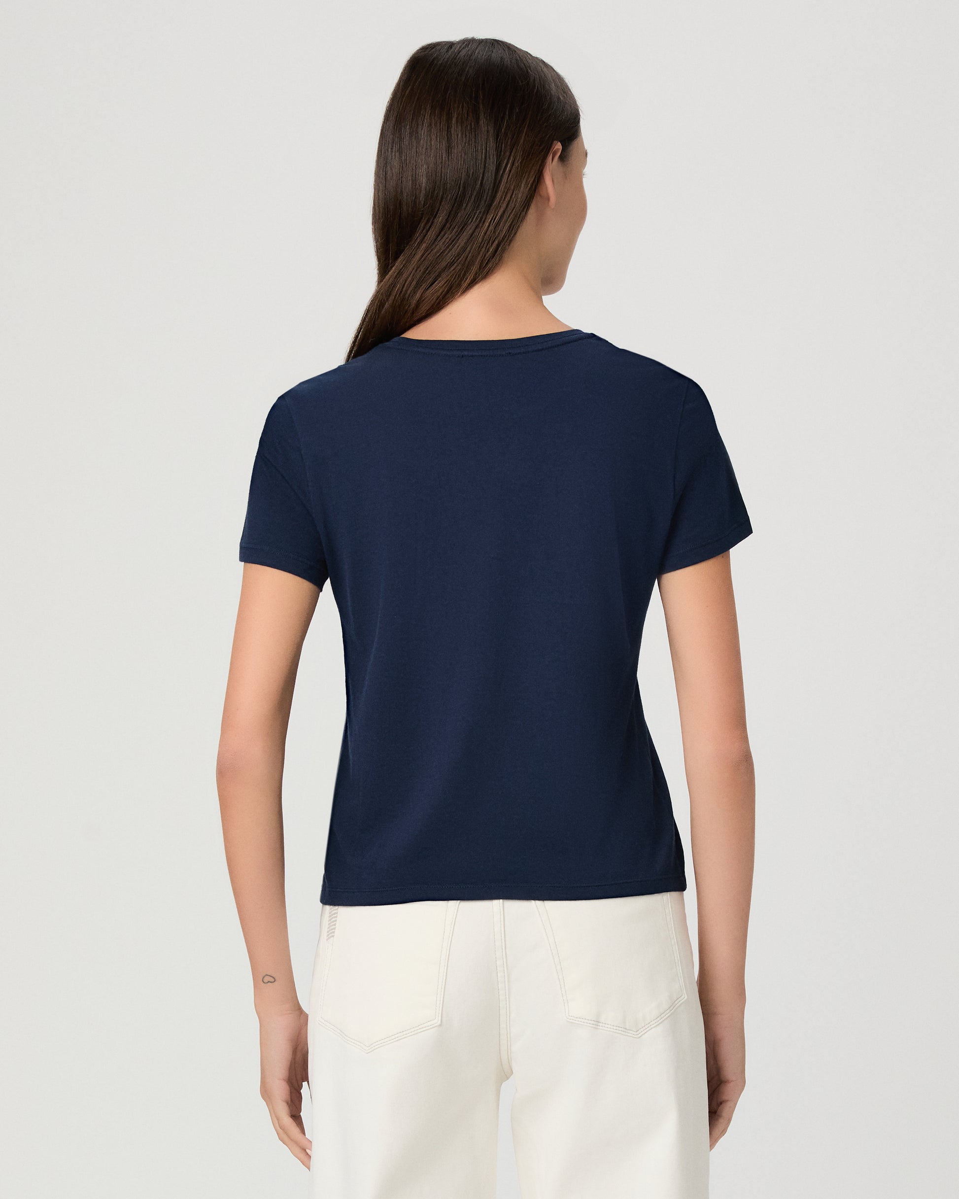 A woman modeling a navy blue short-sleeve crewneck t-shirt paired with off-white pants, shot against a solid light gray background.