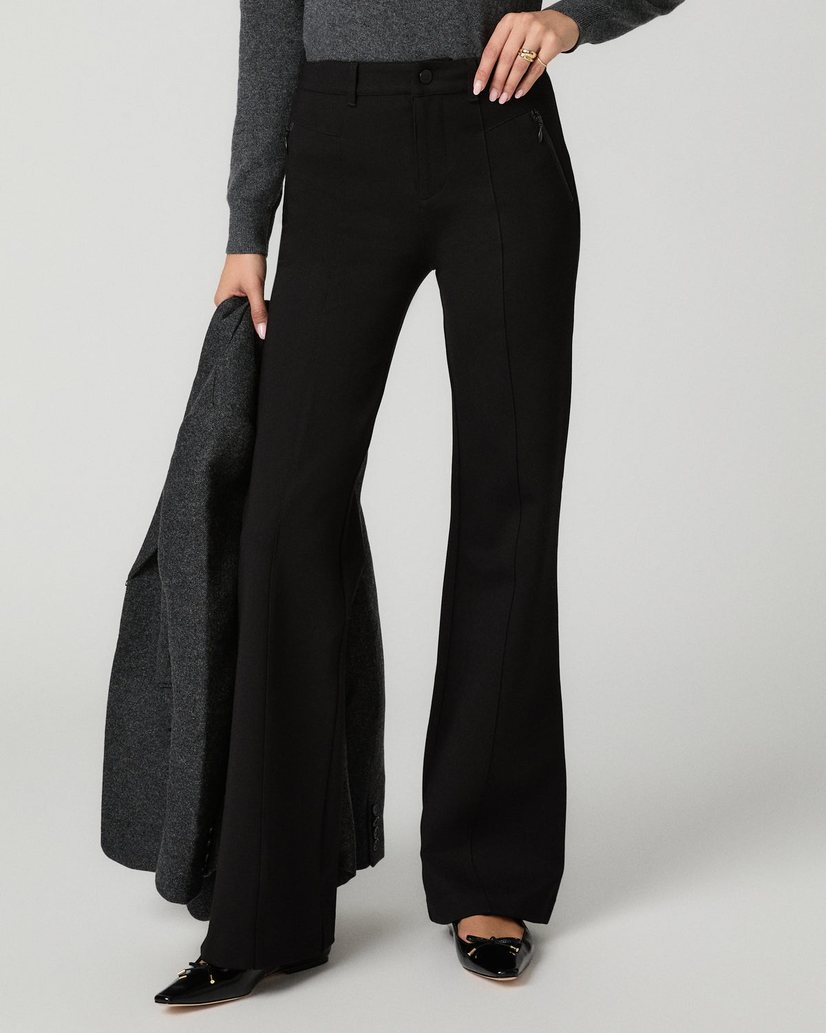 A model wearing black wide-leg trousers, a gray knit sweater, and black patent leather shoes, holding a dark gray coat against a light gray studio background.