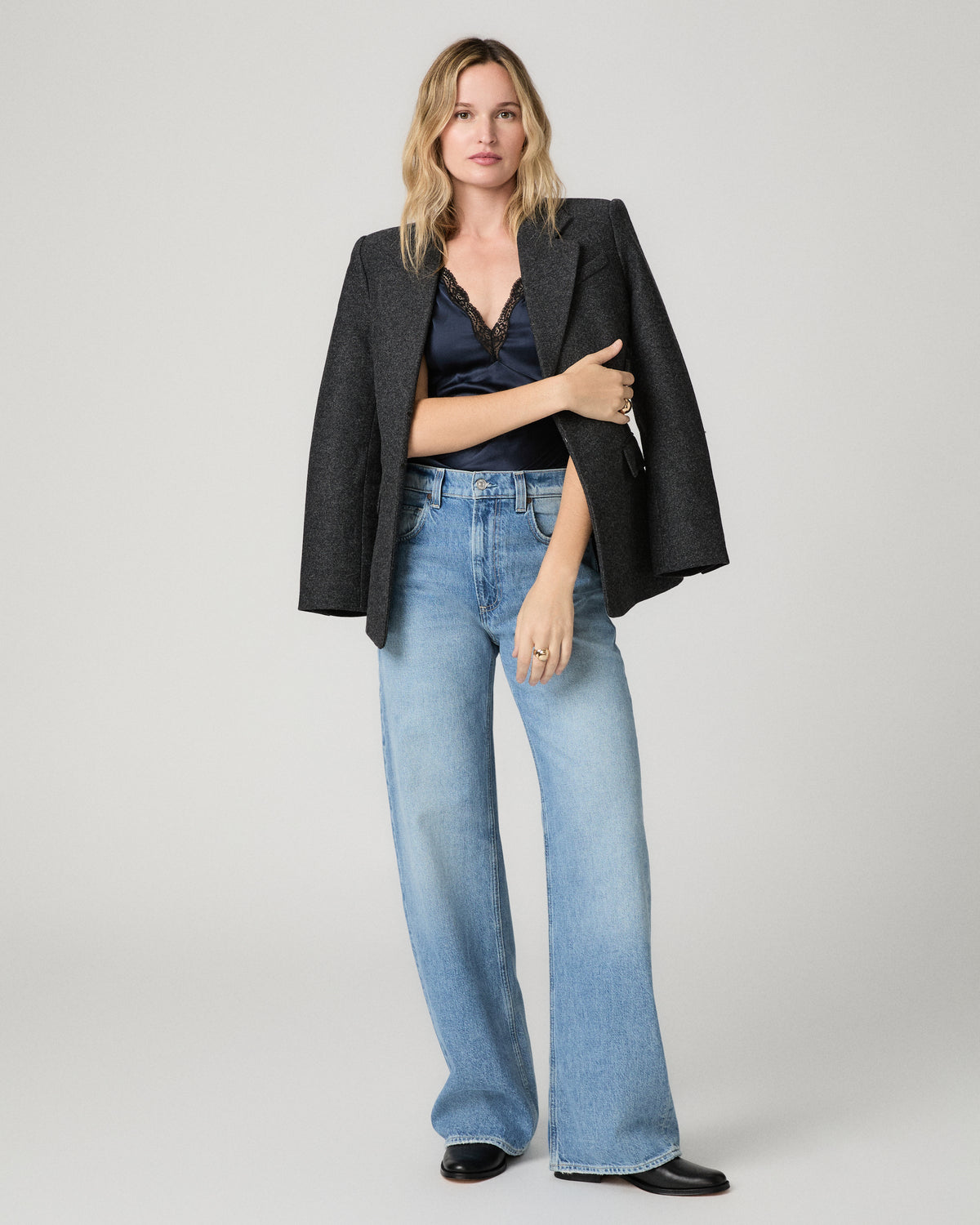 Full-length view of a woman wearing the navy satin Marin Top under a charcoal blazer draped over her shoulders, paired with wide-leg light-wash jeans and black loafers.