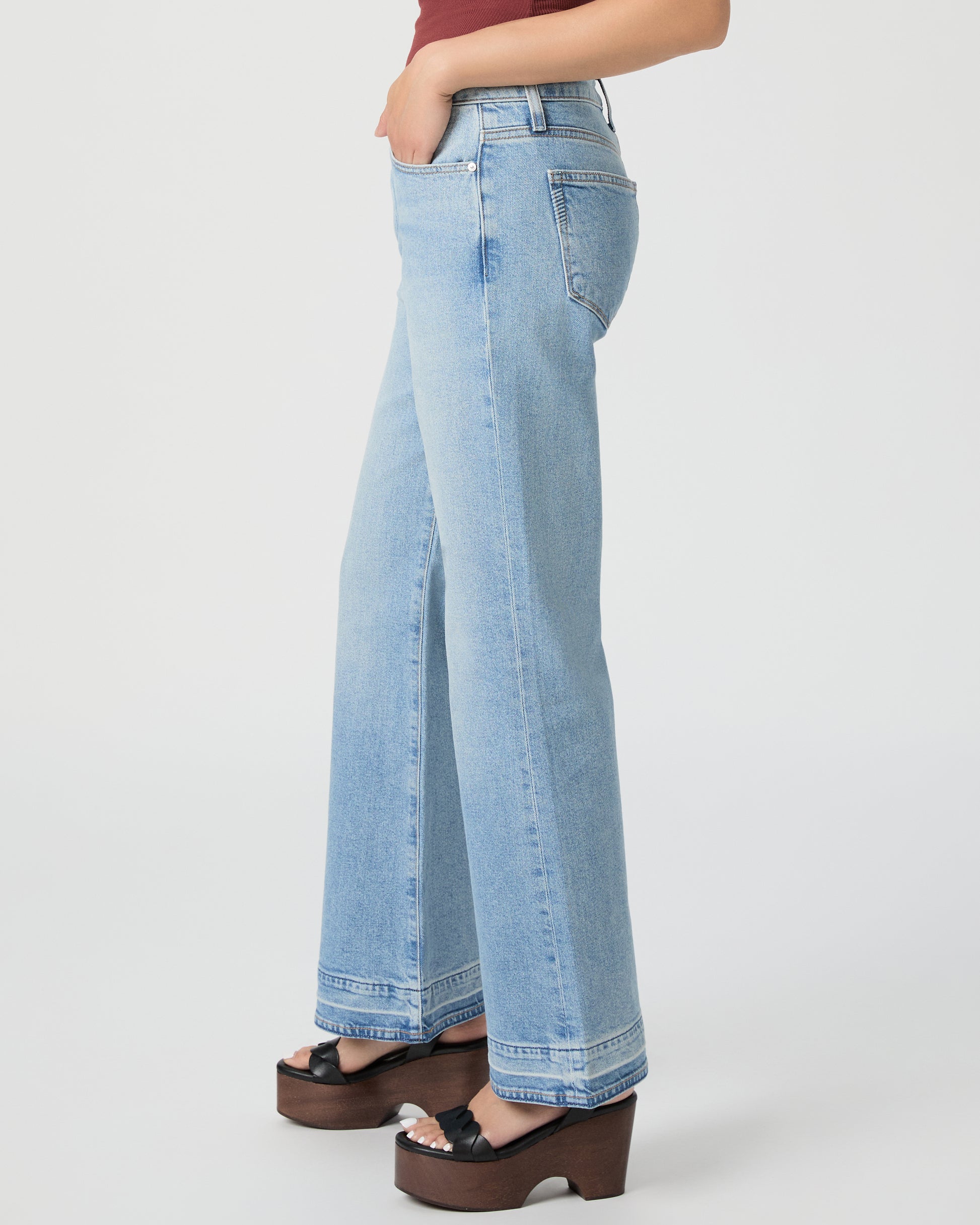 Woman wearing a sleeveless burgundy top and light blue jeans paired with black wedges on a white background