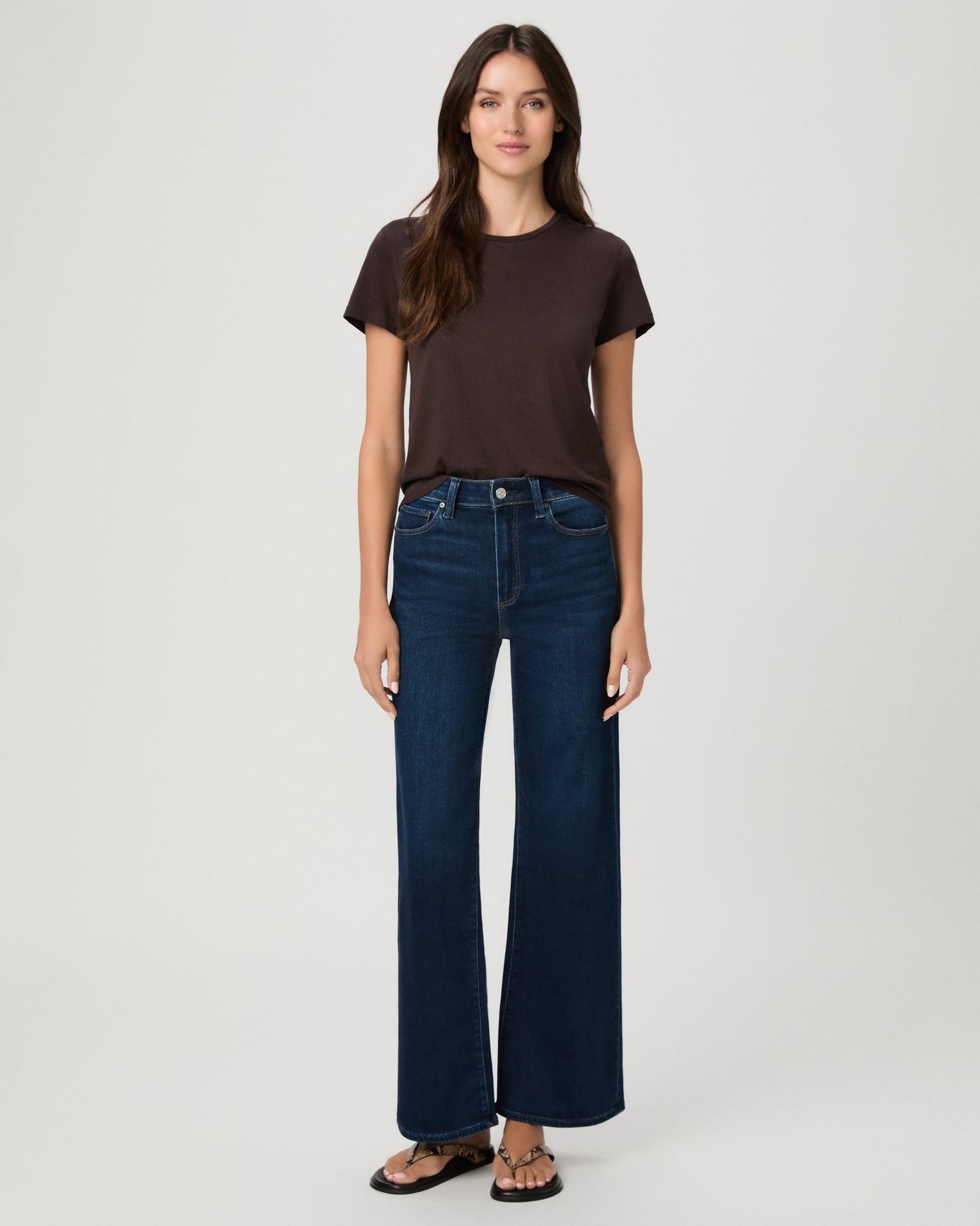 A woman stands against a light gray studio background wearing a dark brown short-sleeve t-shirt, dark blue wide-leg jeans, and brown sandals.