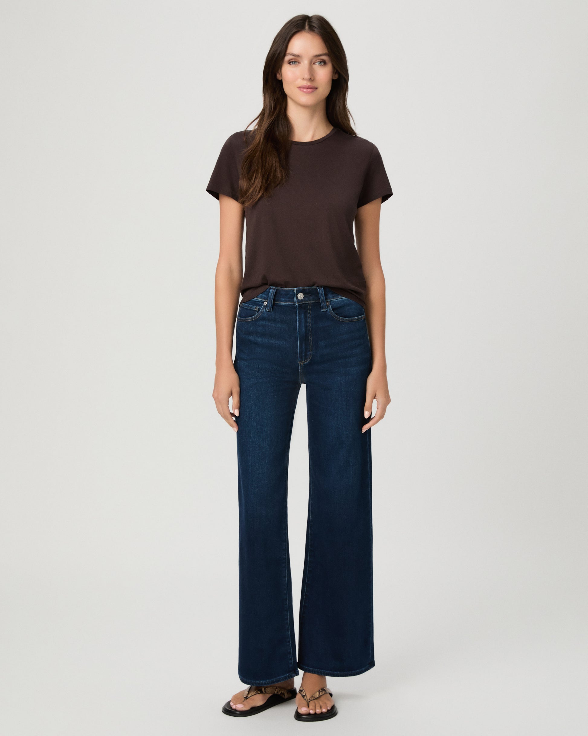 A woman stands against a light gray studio background wearing a dark brown short-sleeve t-shirt, dark blue wide-leg jeans, and brown sandals.