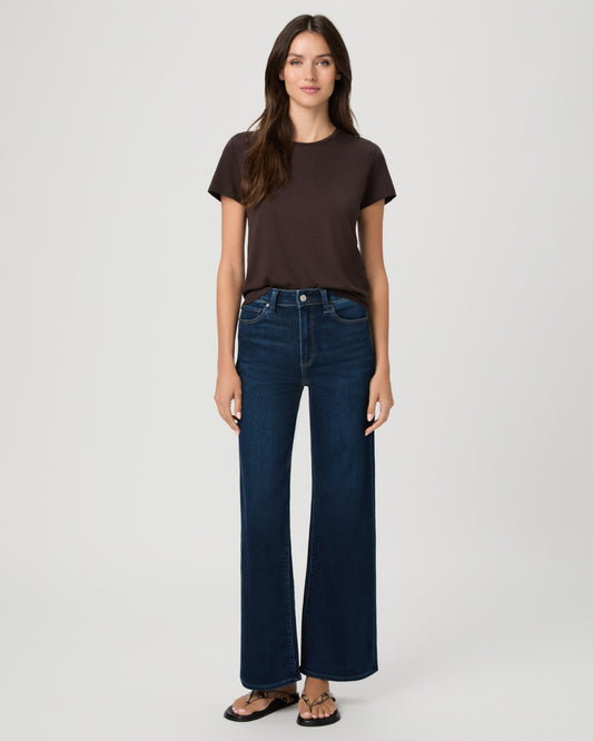 A woman stands against a light gray studio background wearing a dark brown short-sleeve t-shirt, dark blue wide-leg jeans, and brown sandals.
