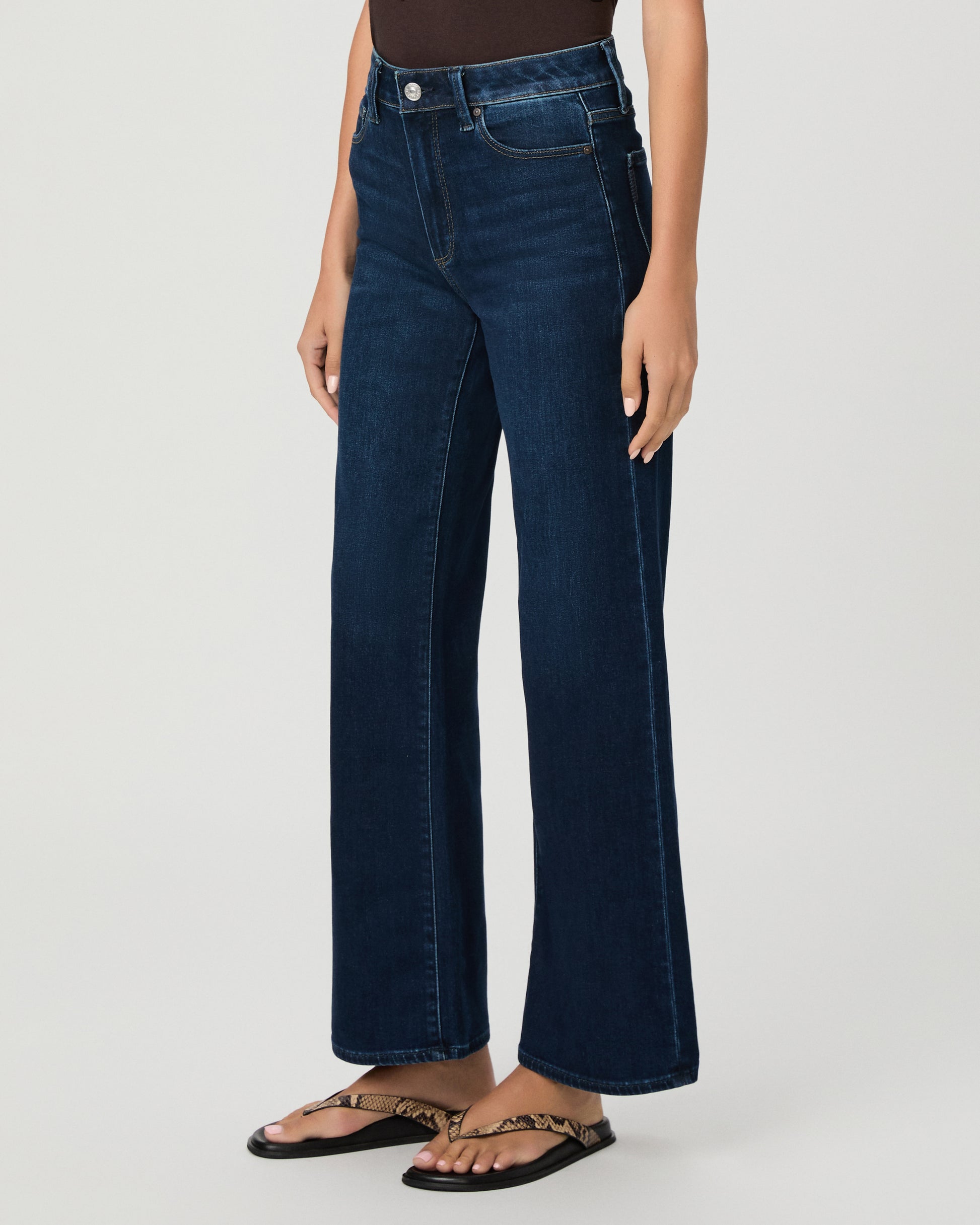 A woman stands against a light gray studio background wearing a dark brown short-sleeve t-shirt, dark blue wide-leg jeans, and brown sandals.