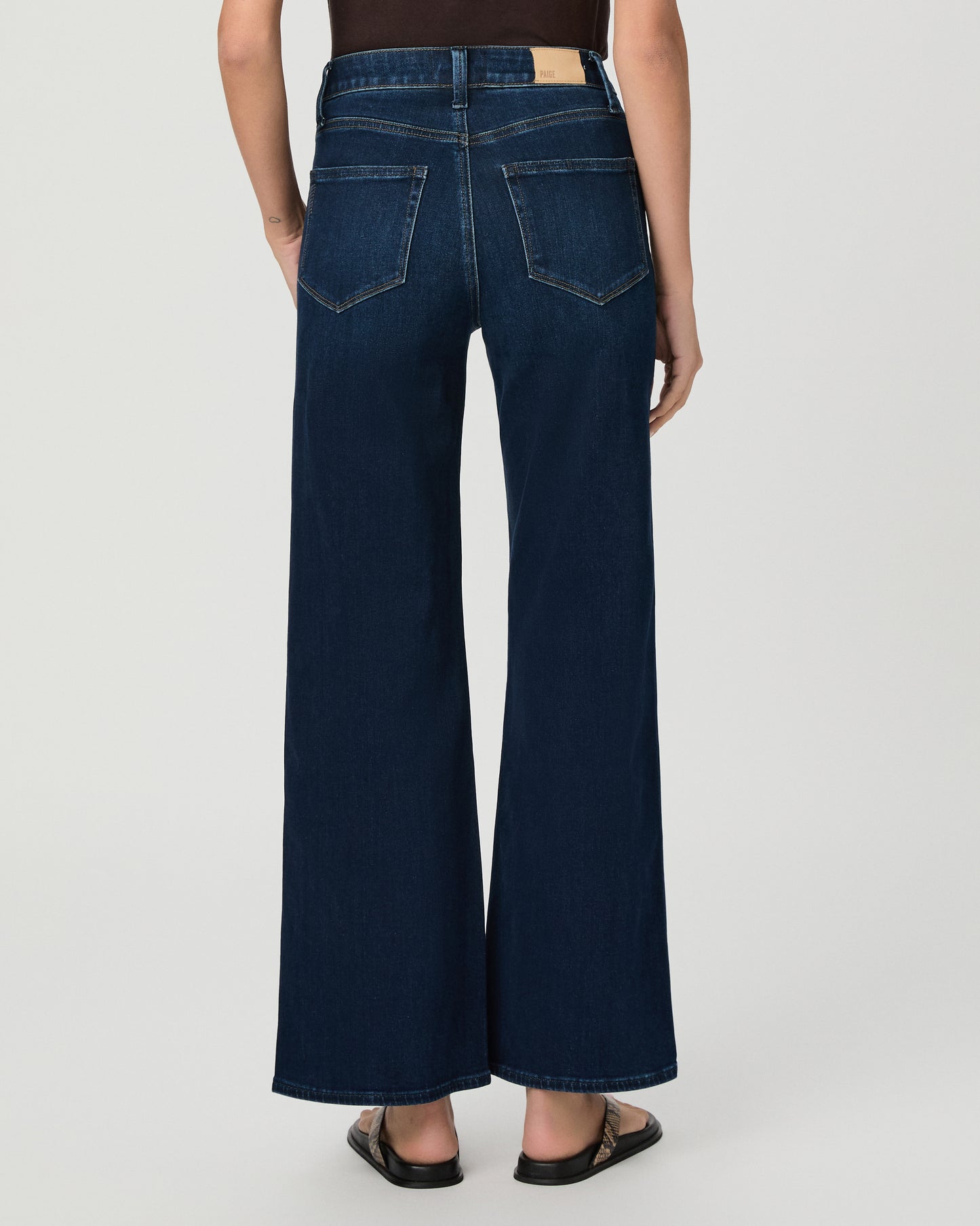 A woman stands against a light gray studio background wearing a dark brown short-sleeve t-shirt, dark blue wide-leg jeans, and brown sandals.