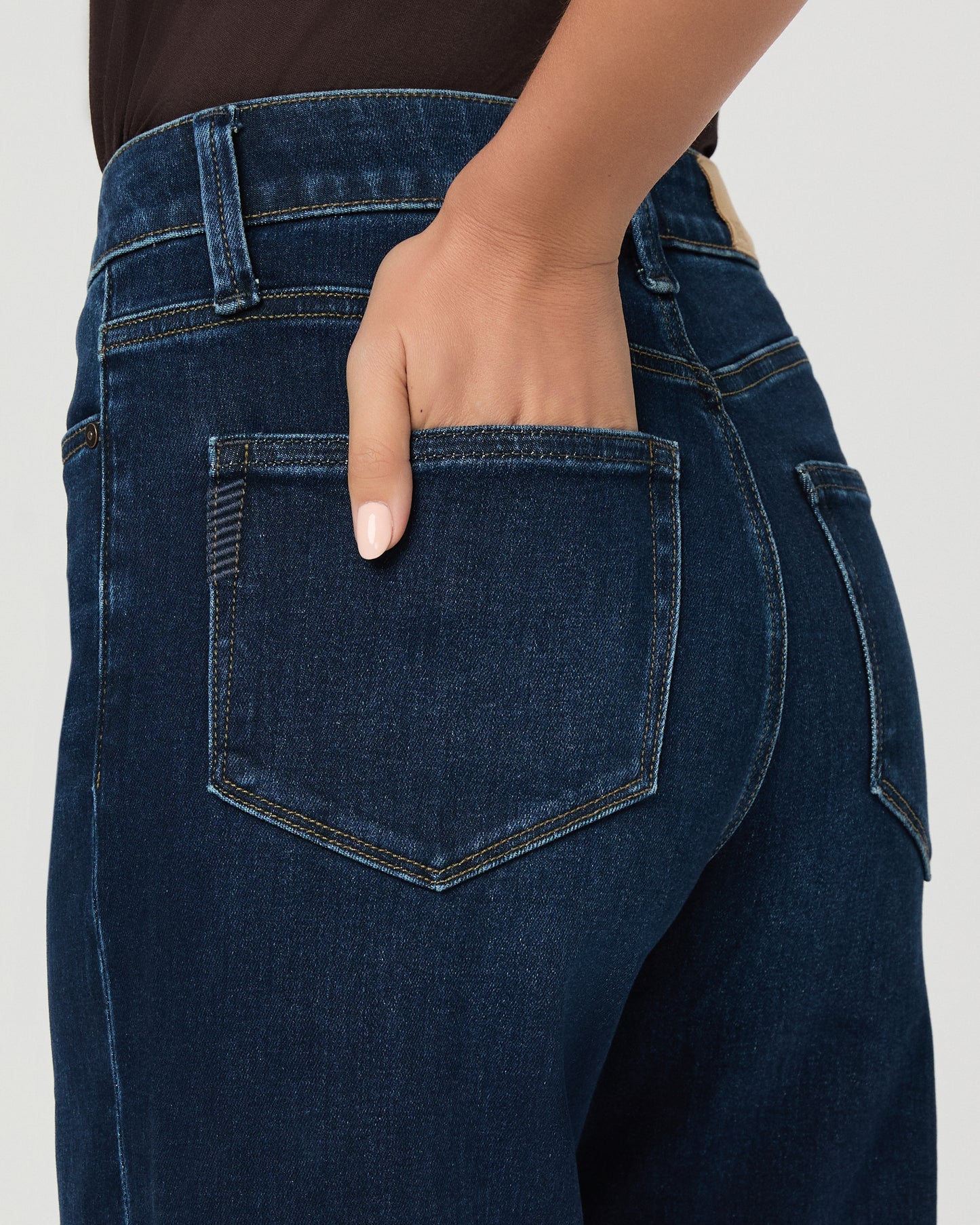 A woman stands against a light gray studio background wearing a dark brown short-sleeve t-shirt, dark blue wide-leg jeans, and brown sandals.