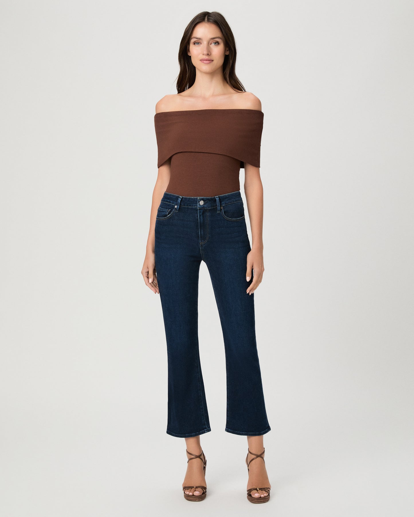 Woman wearing an off the shoulder brown top paired with dark blue jeans and brown wedges against a white background