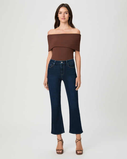 Woman wearing an off the shoulder brown top paired with dark blue jeans and brown wedges against a white background