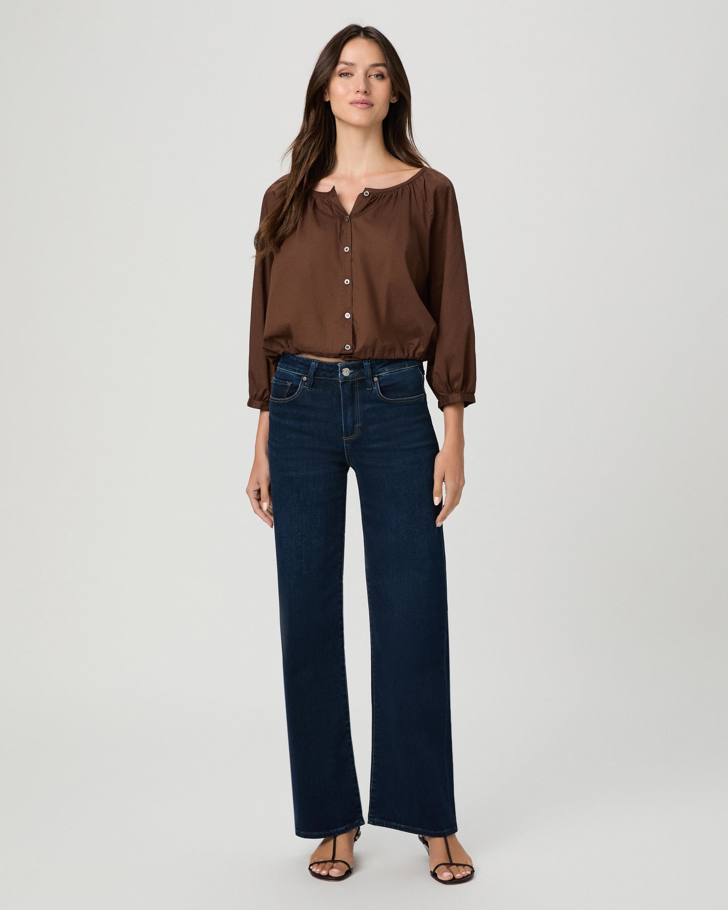 A woman stands against a light gray studio background wearing a brown long-sleeve button-front blouse with a split neckline and dark blue wide-leg jeans.