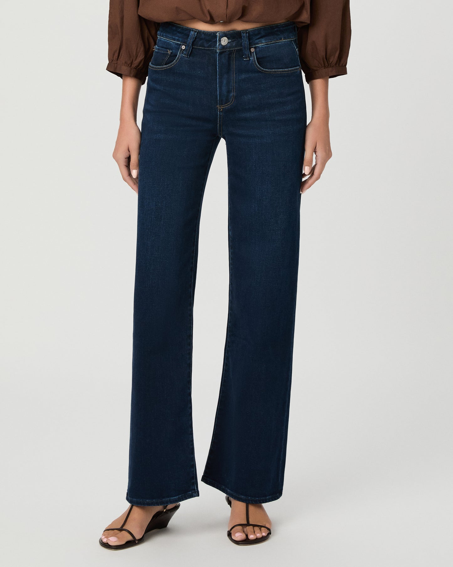 A woman stands against a light gray studio background wearing a brown long-sleeve button-front blouse with a split neckline and dark blue wide-leg jeans.