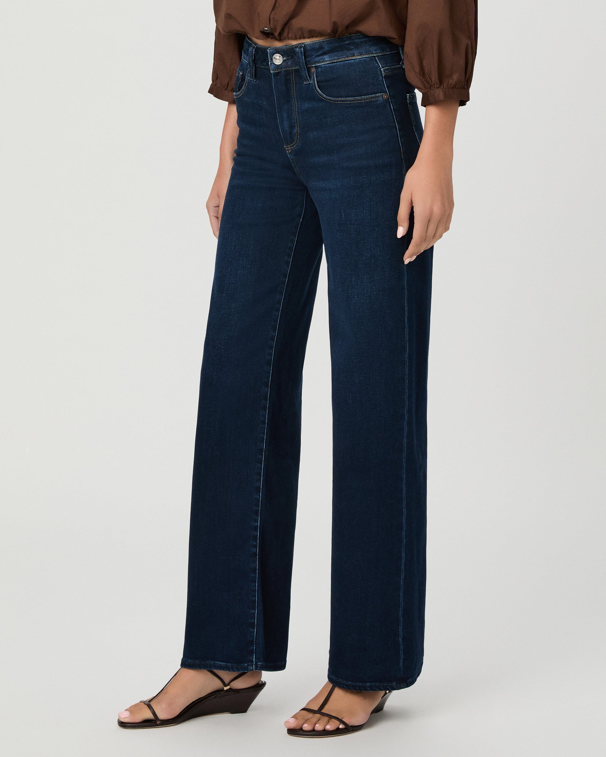 A woman stands against a light gray studio background wearing a brown long-sleeve button-front blouse with a split neckline and dark blue wide-leg jeans.