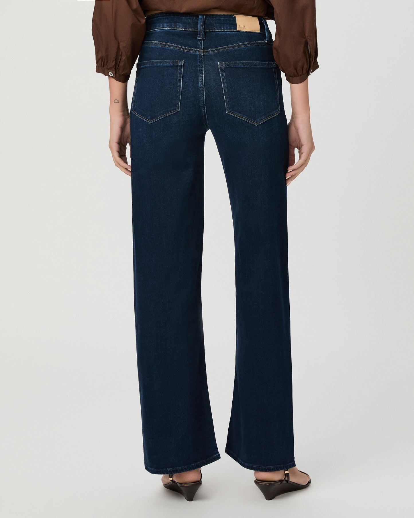 A woman stands against a light gray studio background wearing a brown long-sleeve button-front blouse with a split neckline and dark blue wide-leg jeans.