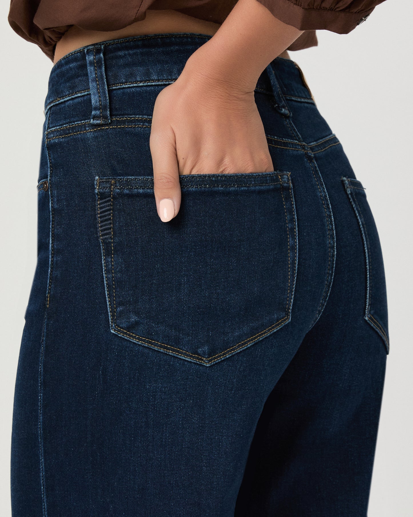 A woman stands against a light gray studio background wearing a brown long-sleeve button-front blouse with a split neckline and dark blue wide-leg jeans.