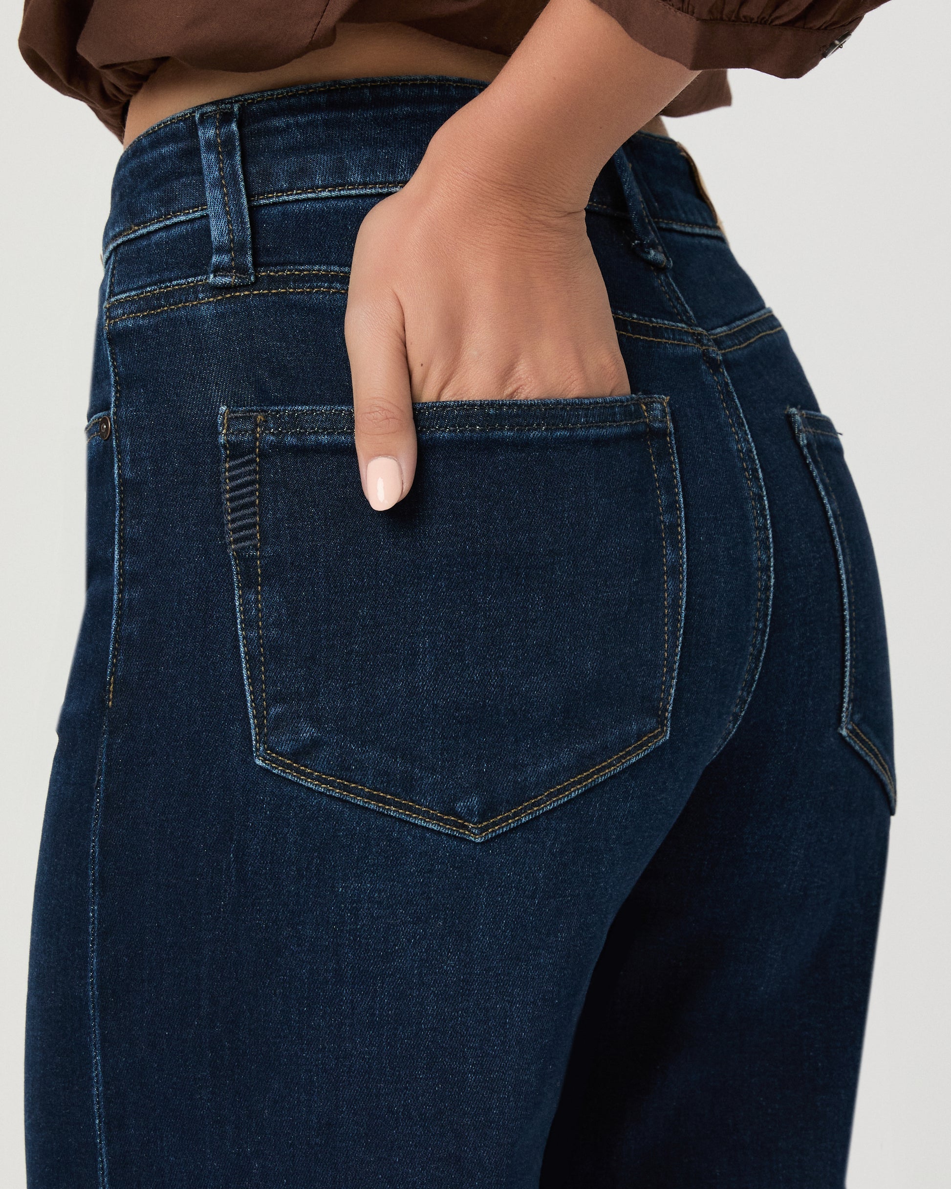 A woman stands against a light gray studio background wearing a brown long-sleeve button-front blouse with a split neckline and dark blue wide-leg jeans.