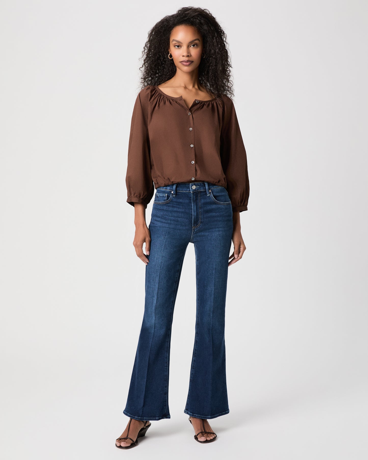 A woman modeling a brown long-sleeve button-front blouse with gathered shoulders and dark blue flare-leg jeans against a light white background.
