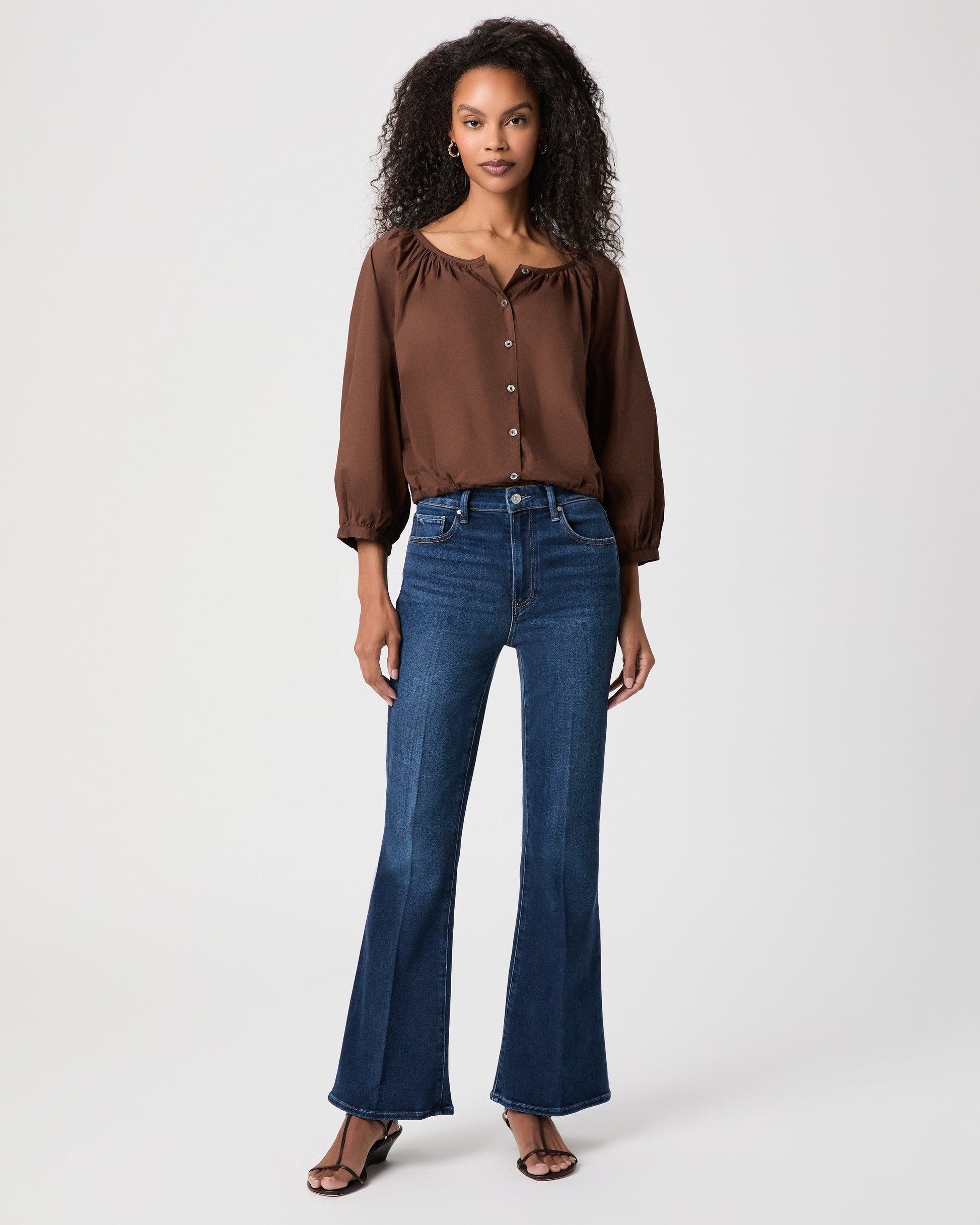 A woman modeling a brown long-sleeve button-front blouse with gathered shoulders and dark blue flare-leg jeans against a light white background.