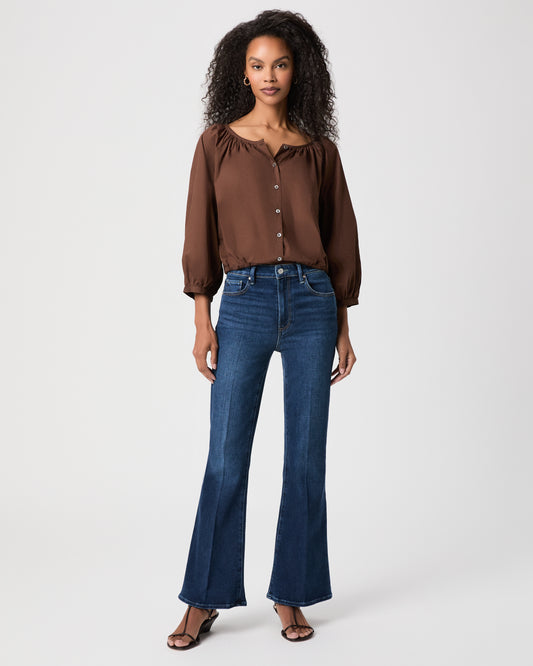 A woman modeling a brown long-sleeve button-front blouse with gathered shoulders and dark blue flare-leg jeans against a light white background.