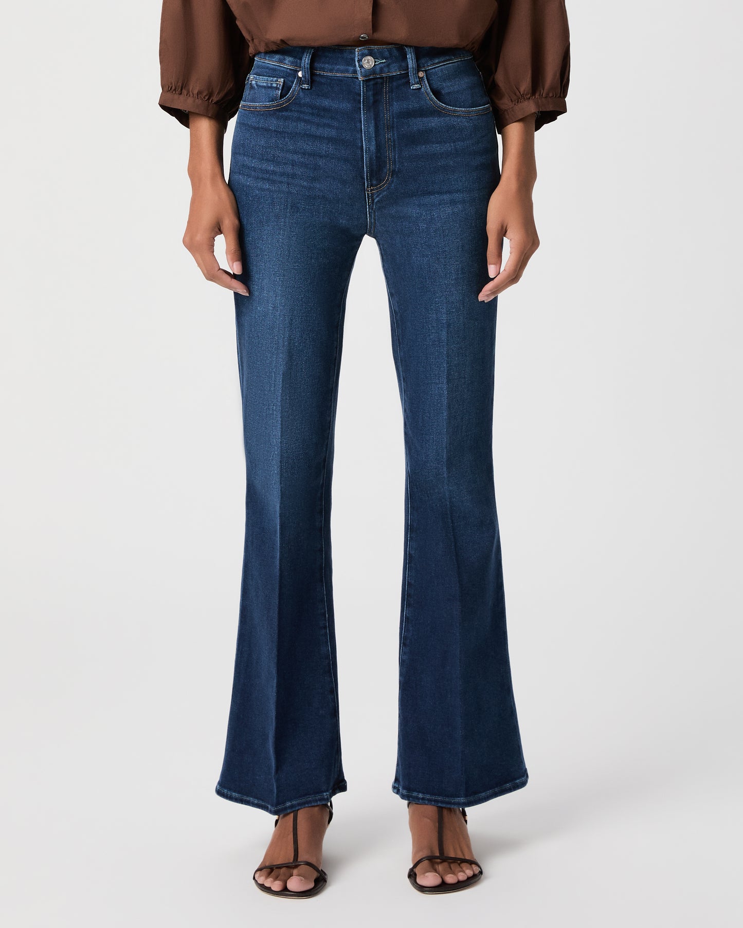 A woman modeling a brown long-sleeve button-front blouse with gathered shoulders and dark blue flare-leg jeans against a light white background.