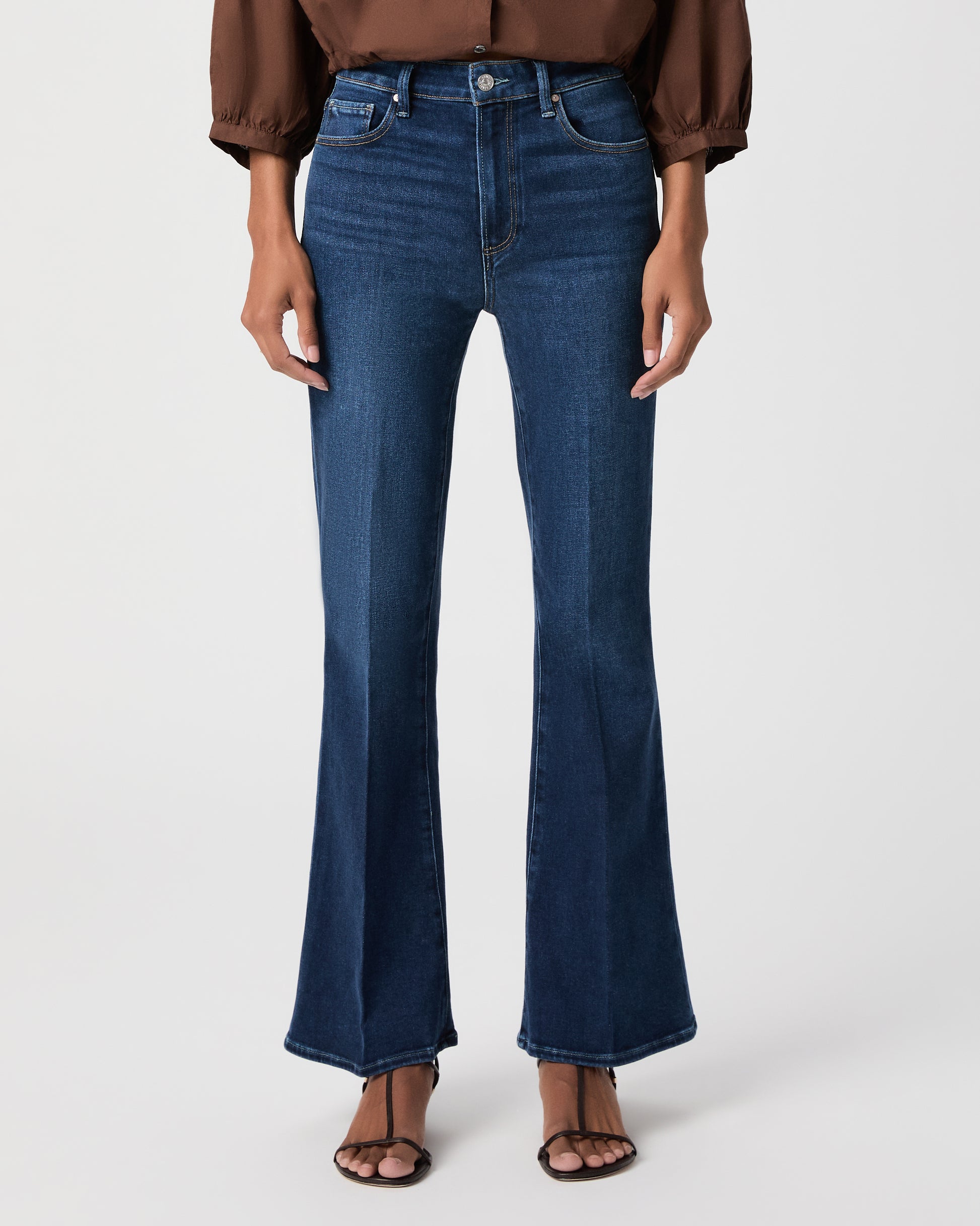 A woman modeling a brown long-sleeve button-front blouse with gathered shoulders and dark blue flare-leg jeans against a light white background.