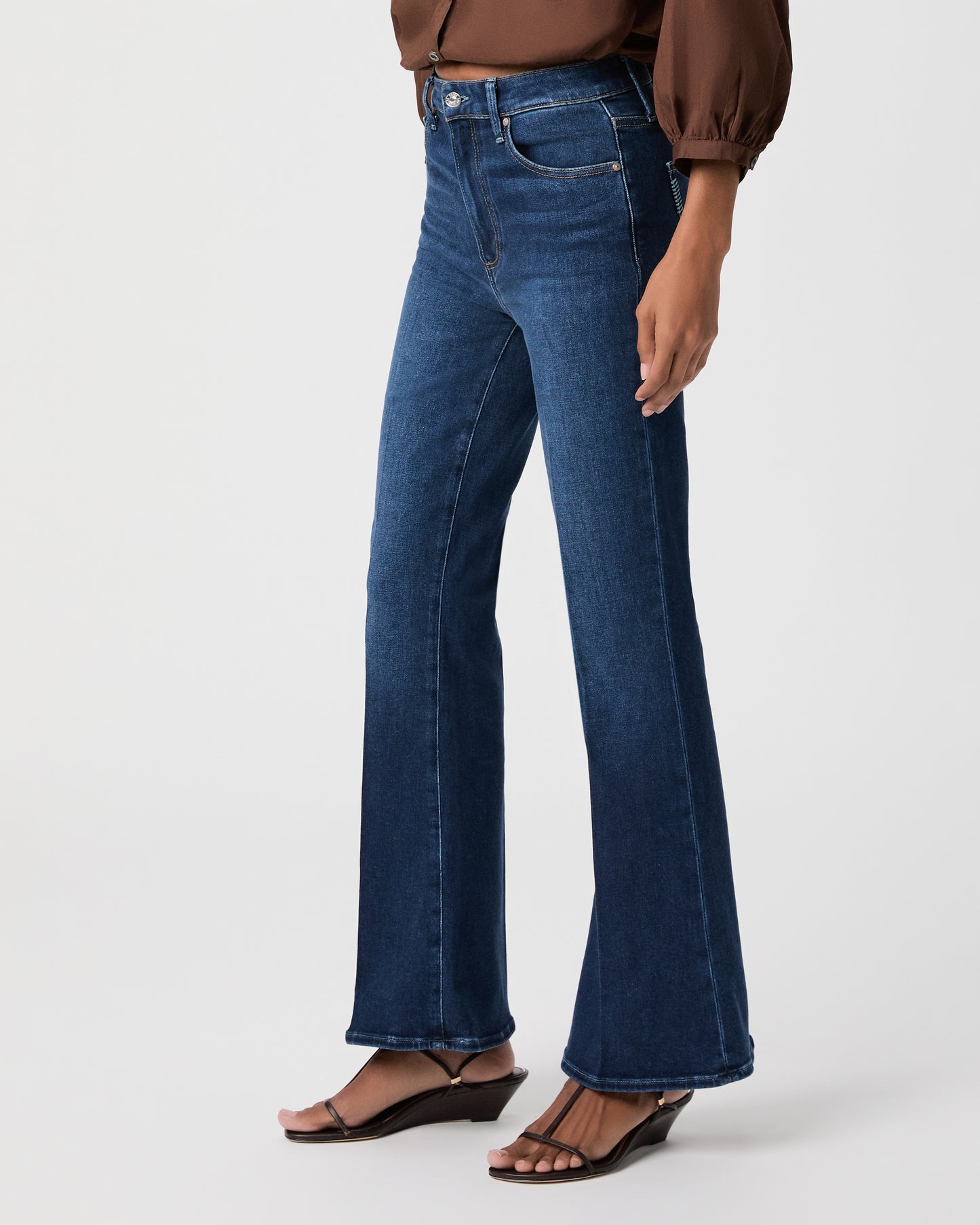A woman modeling a brown long-sleeve button-front blouse with gathered shoulders and dark blue flare-leg jeans against a light white background.