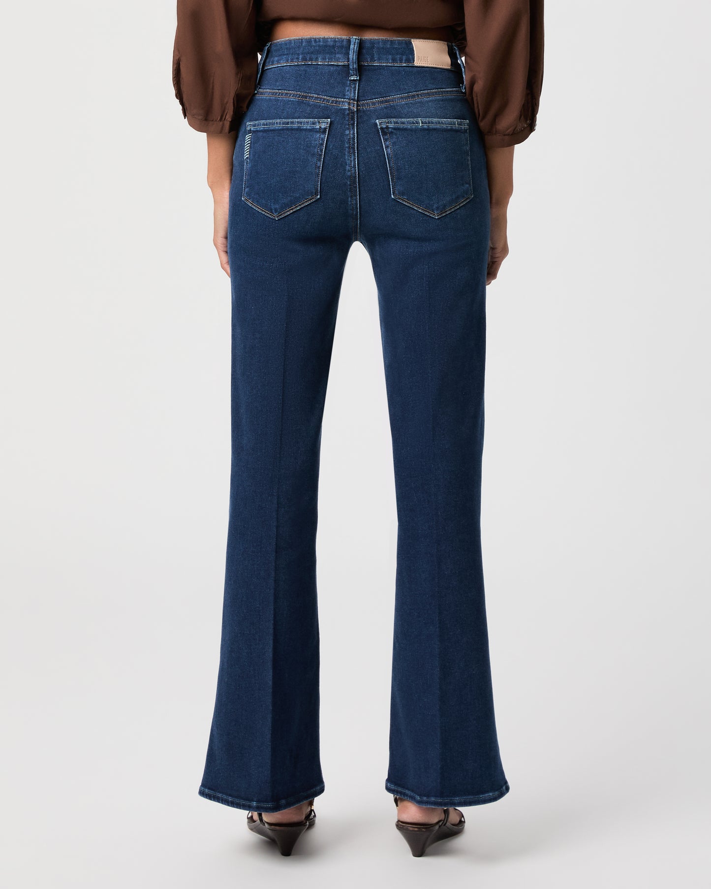 A woman modeling a brown long-sleeve button-front blouse with gathered shoulders and dark blue flare-leg jeans against a light white background.