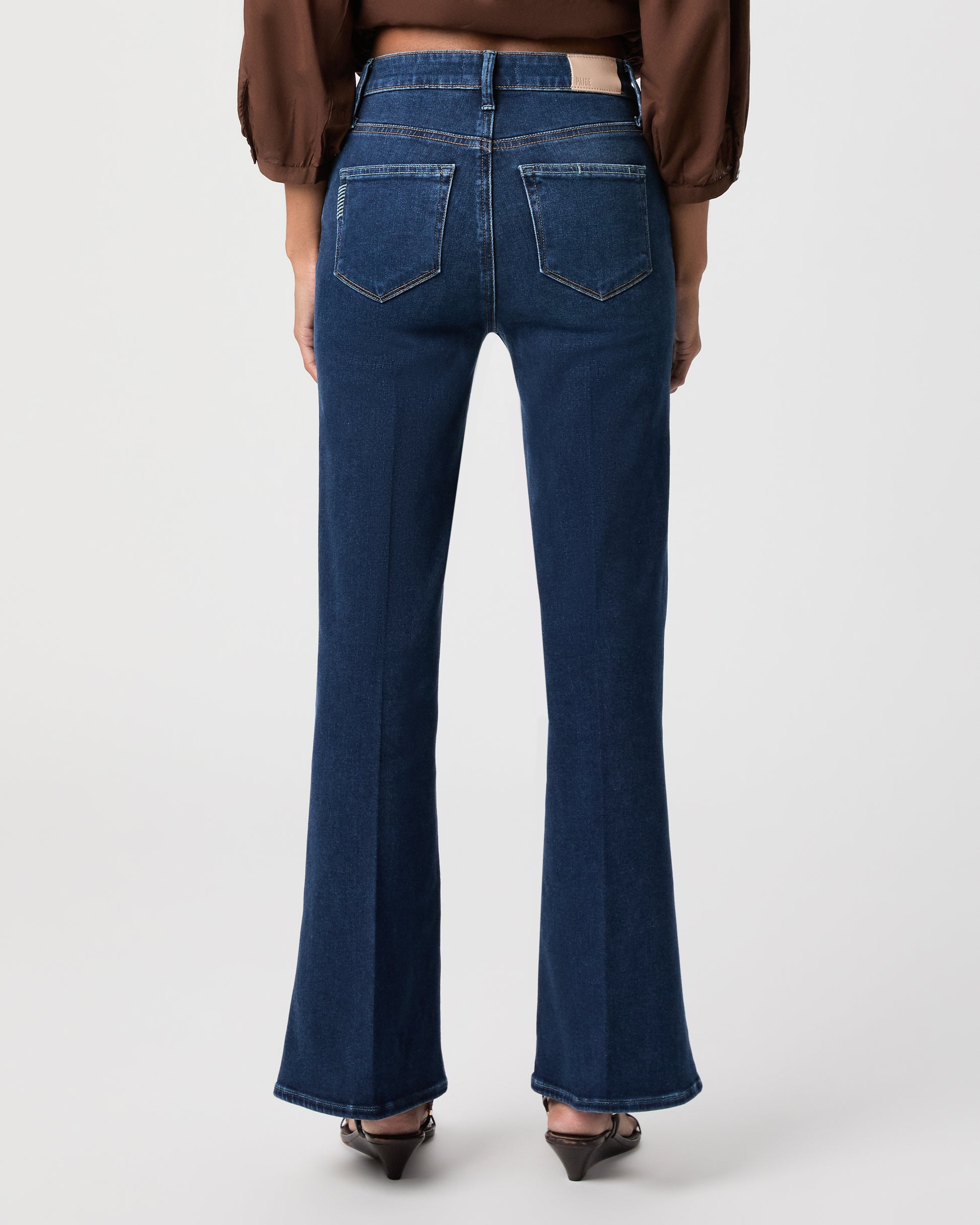 A woman modeling a brown long-sleeve button-front blouse with gathered shoulders and dark blue flare-leg jeans against a light white background.