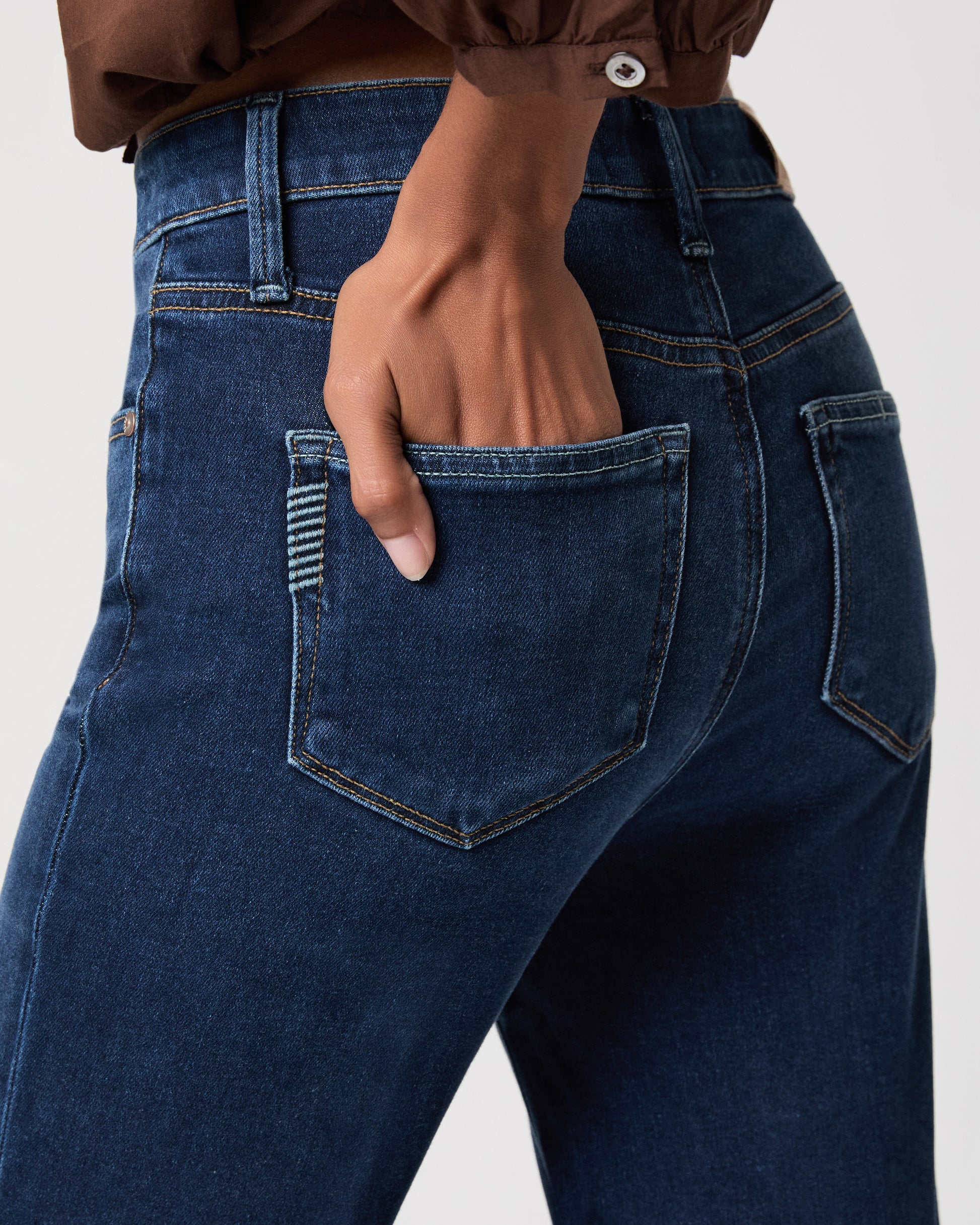 A woman modeling a brown long-sleeve button-front blouse with gathered shoulders and dark blue flare-leg jeans against a light white background.