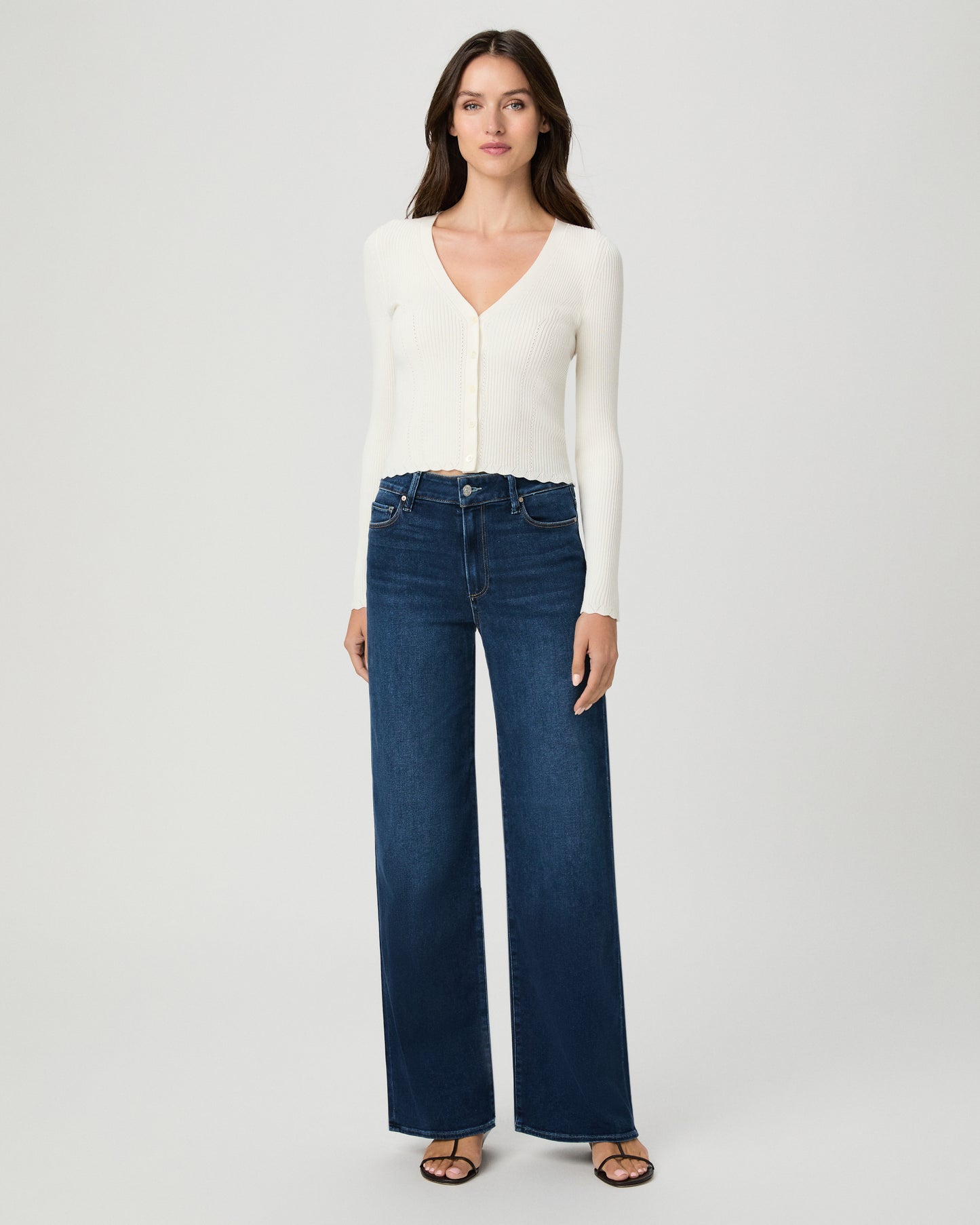 A woman modeling a cream long-sleeve button-front cardigan and dark blue wide-leg jeans against a light gray studio background.