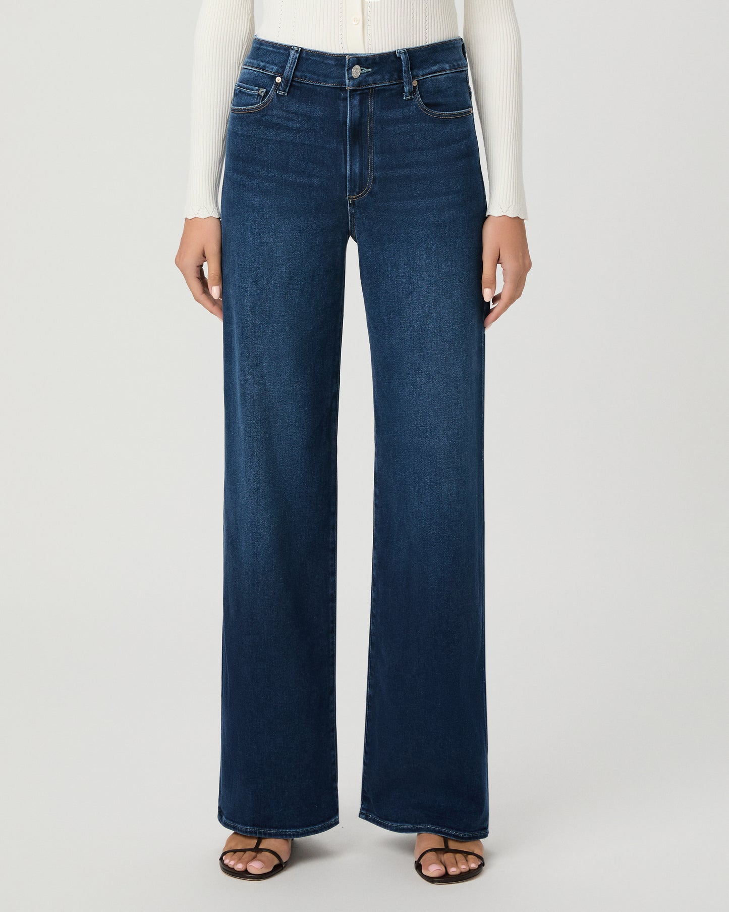 A woman modeling a cream long-sleeve button-front cardigan and dark blue wide-leg jeans against a light gray studio background.