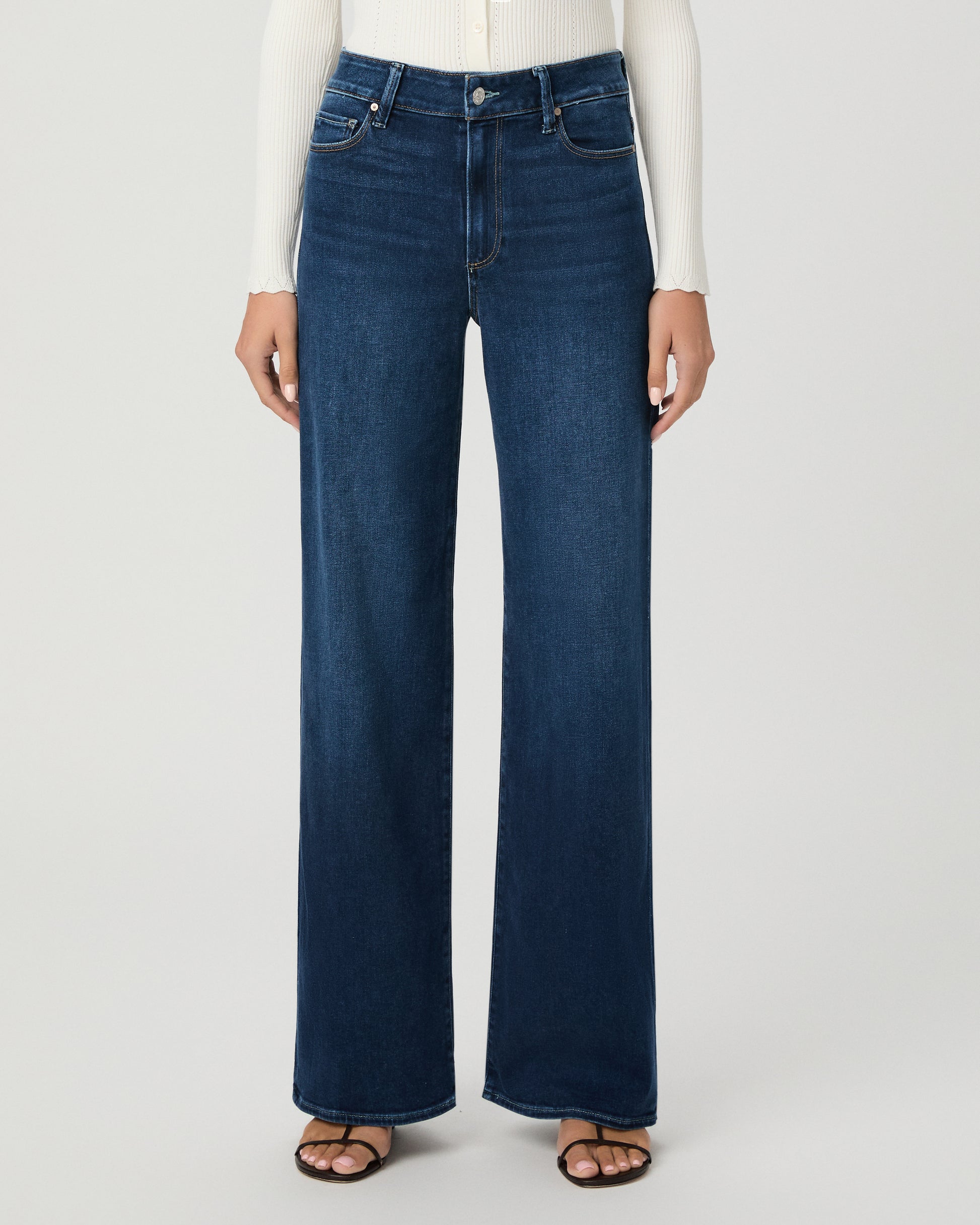 A woman modeling a cream long-sleeve button-front cardigan and dark blue wide-leg jeans against a light gray studio background.