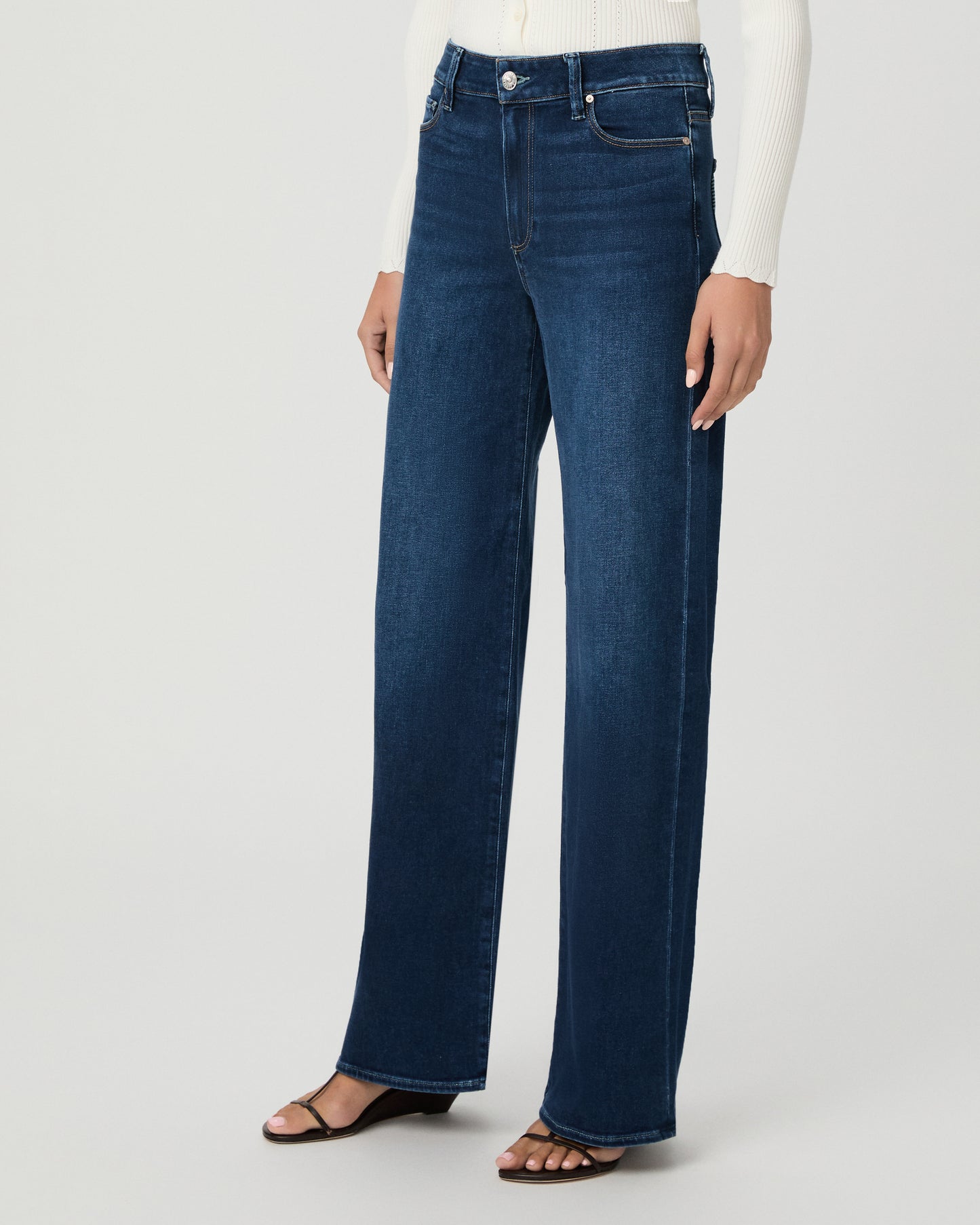 A woman modeling a cream long-sleeve button-front cardigan and dark blue wide-leg jeans against a light gray studio background.