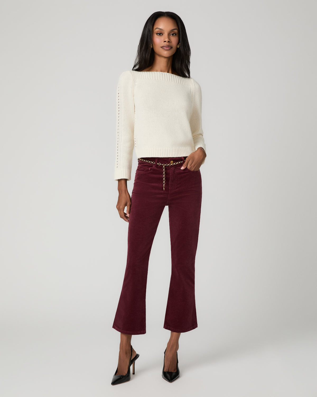 Full-length front view of a woman wearing an ivory ribbed sweater and burgundy corduroy cropped bootcut jeans with button-front detail, paired with black pointed-toe heels.