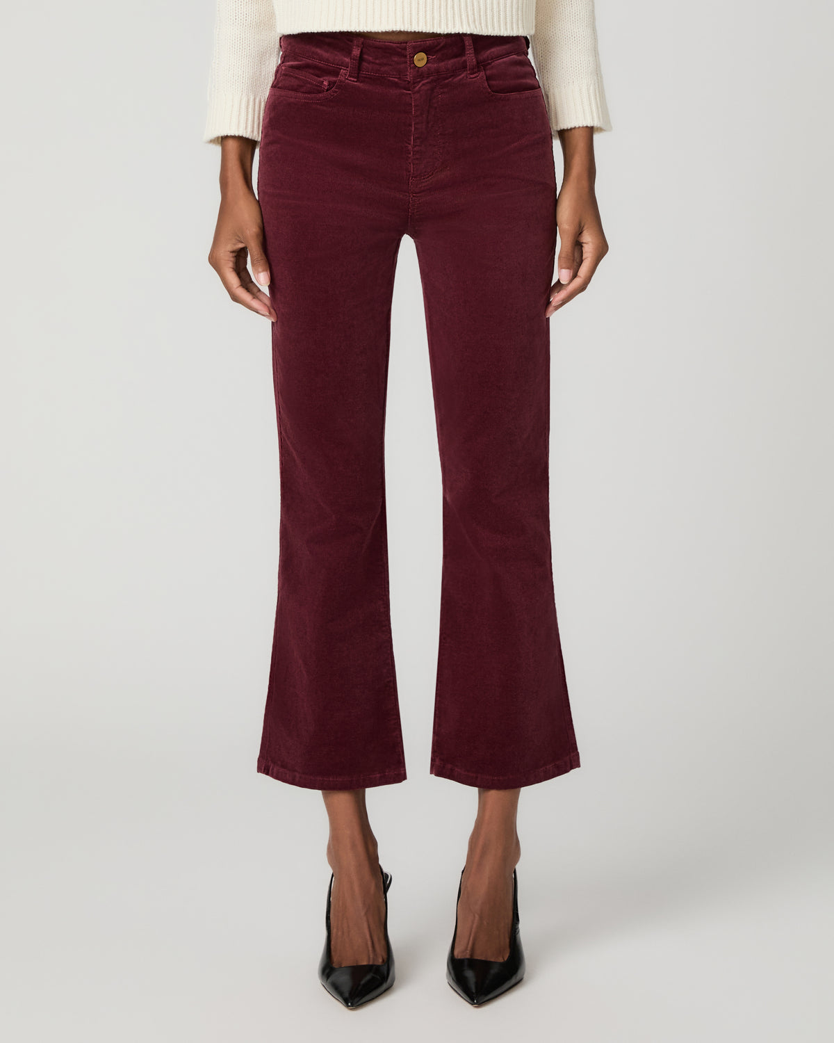 Close-up front view of burgundy corduroy bootcut jeans with a button fly, styled with an ivory sweater and black pointed-toe heels.