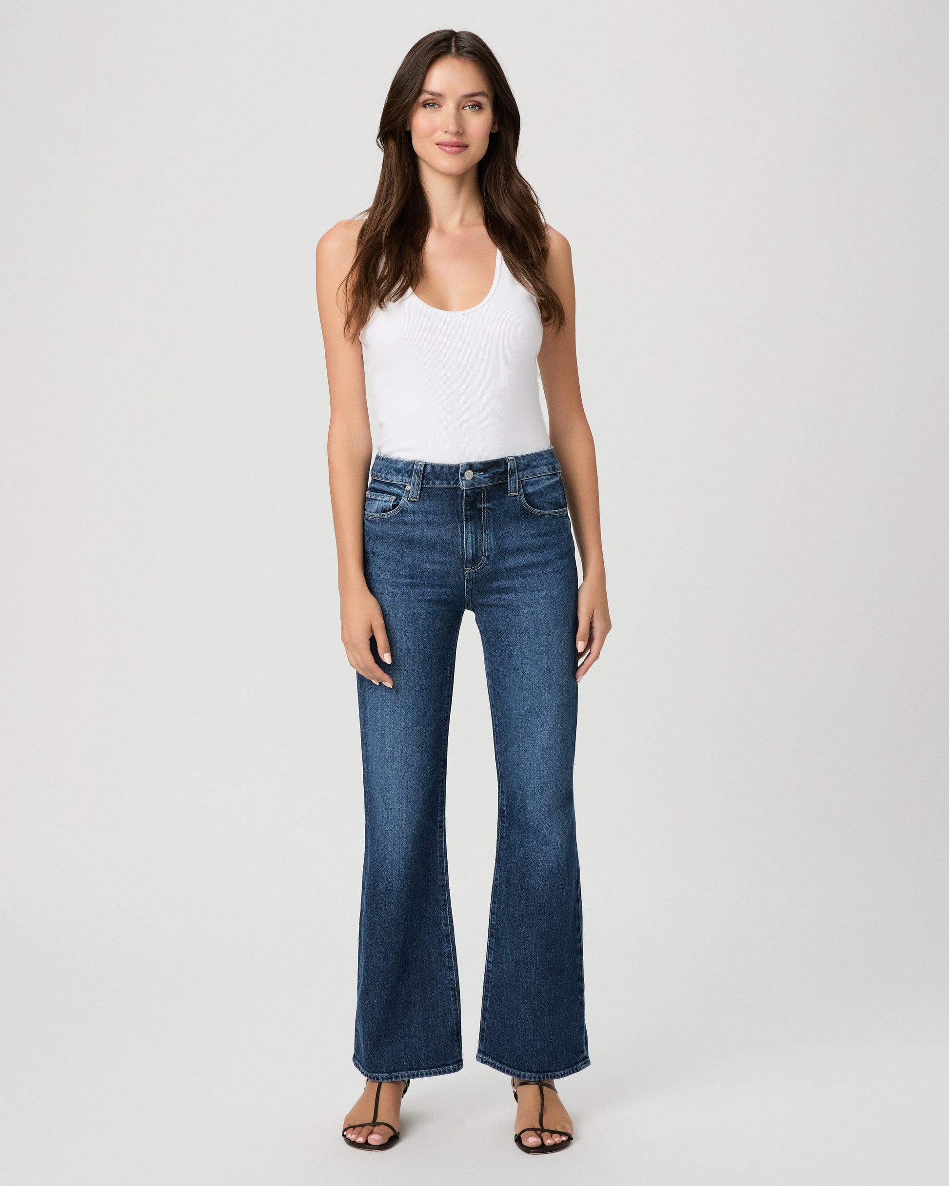A woman modeling a pair of dark blue high-waisted flare jeans with a white sleeveless top, standing against a light gray studio background.
