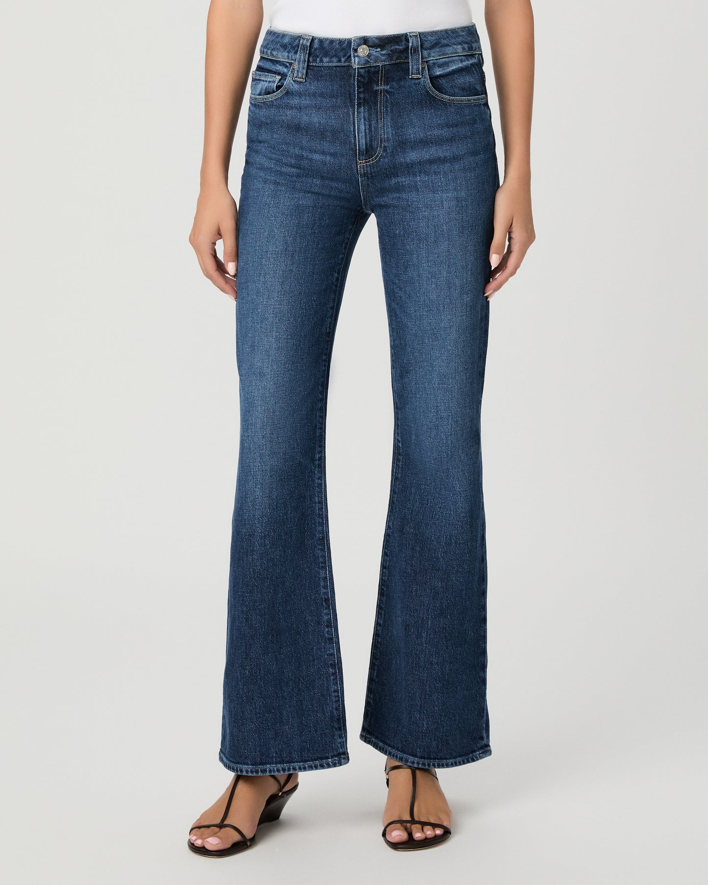A woman modeling a pair of dark blue high-waisted flare jeans with a white sleeveless top, standing against a light gray studio background.
