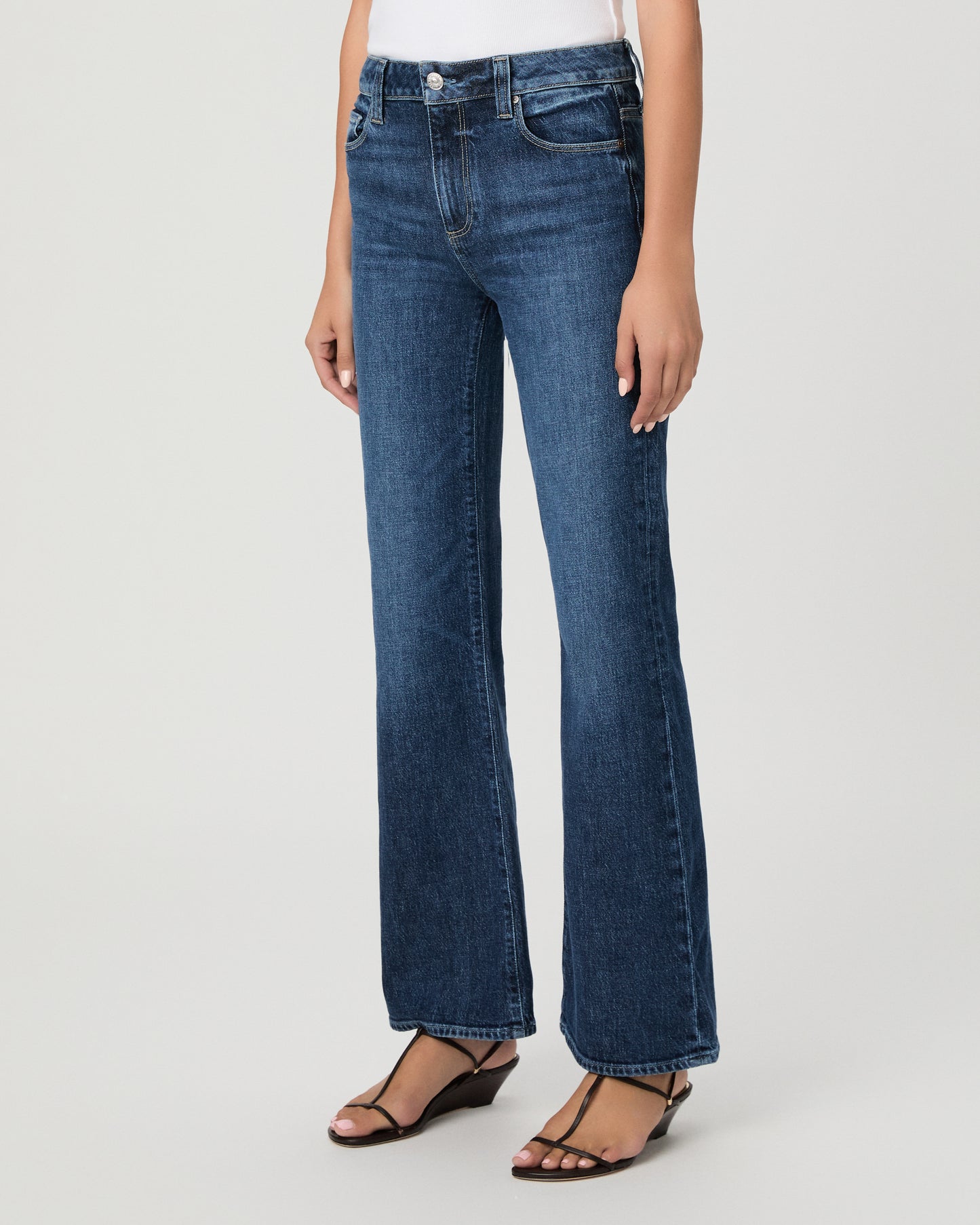 A woman modeling a pair of dark blue high-waisted flare jeans with a white sleeveless top, standing against a light gray studio background.