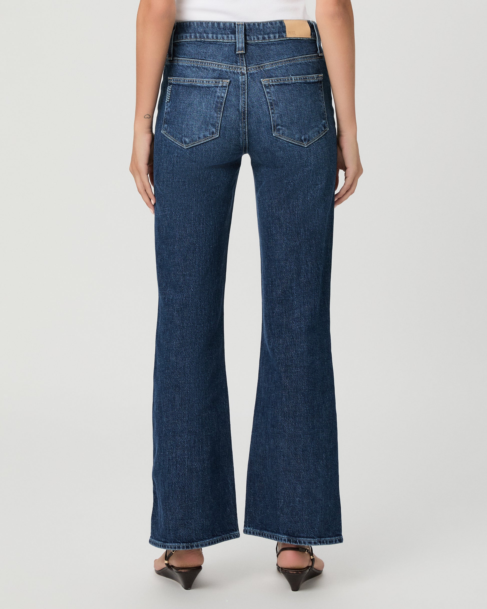 A woman modeling a pair of dark blue high-waisted flare jeans with a white sleeveless top, standing against a light gray studio background.